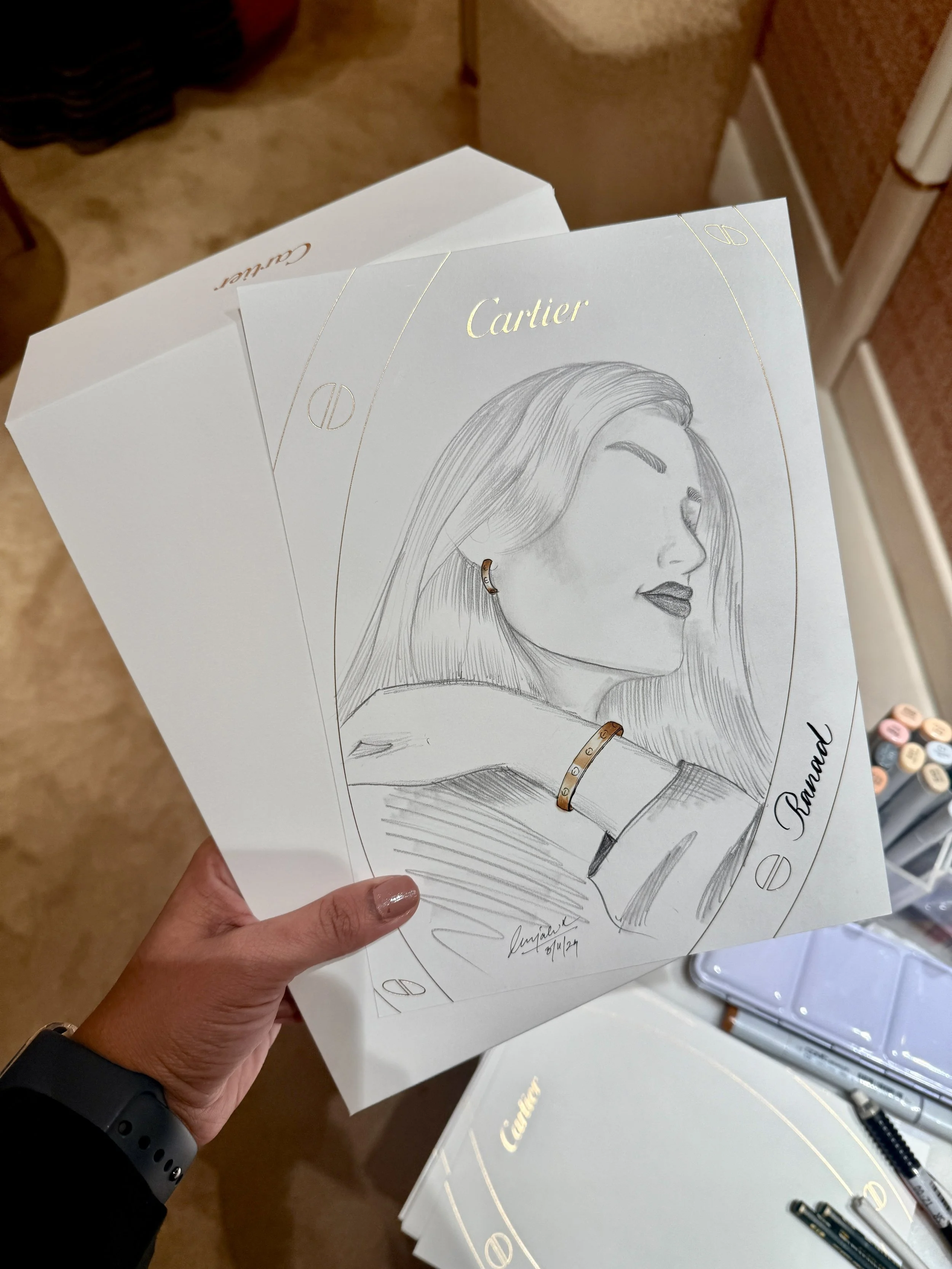 A hand holding a white paper with a sketch of a woman wearing a Cartier bracelet and earring, with a Cartier box and pens on a desk in the background.