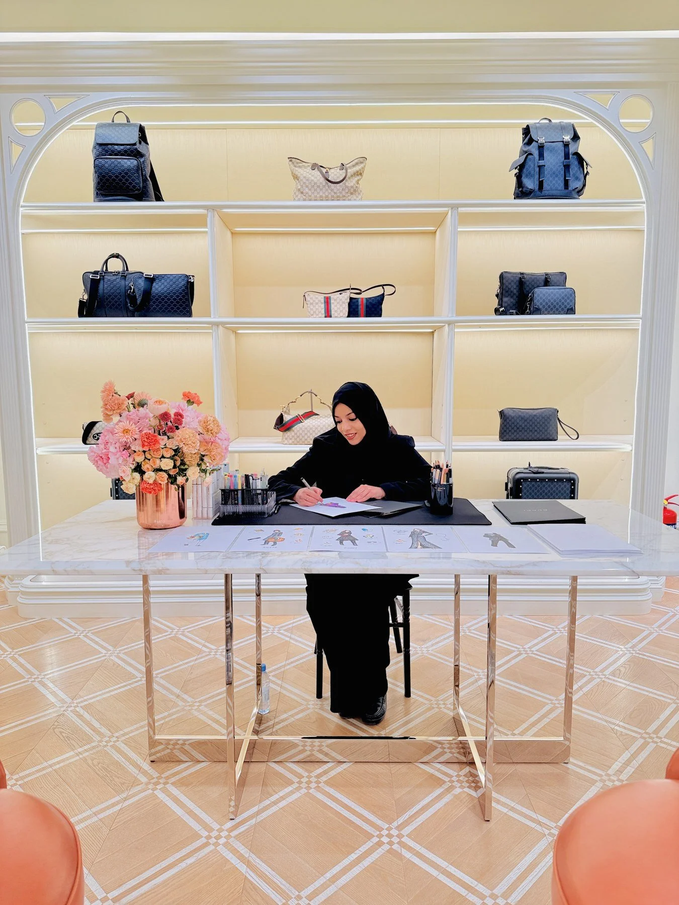 A woman sitting at a table, drawing fashion sketches, with a large vase of pink and peach flowers and organized markers on the table. Behind her is a display shelf with handbags and accessories.