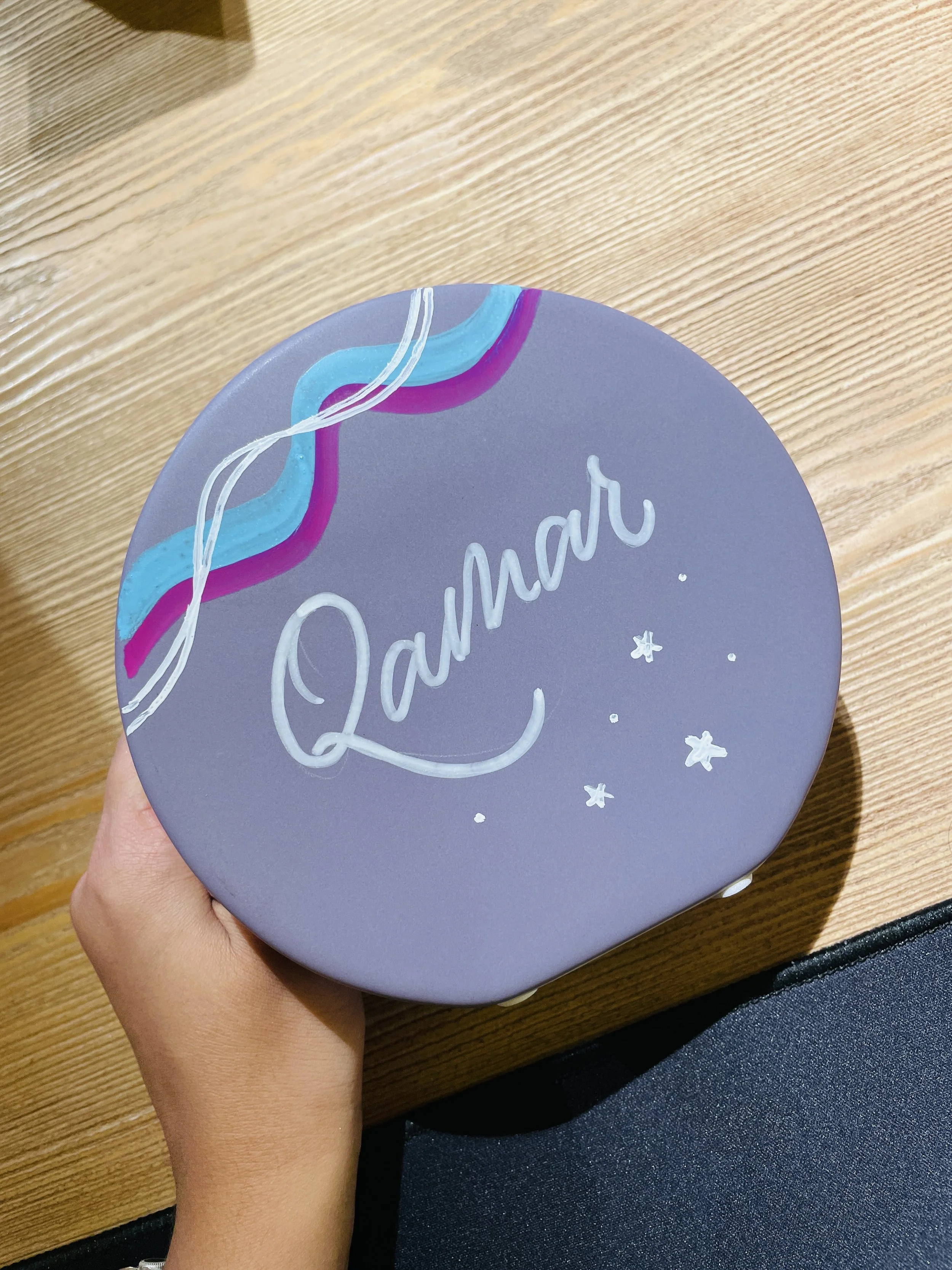Close-up of a person holding a round purple object with the word 'Lunar' written on it, decorated with a rainbow and star designs, on a wooden surface.