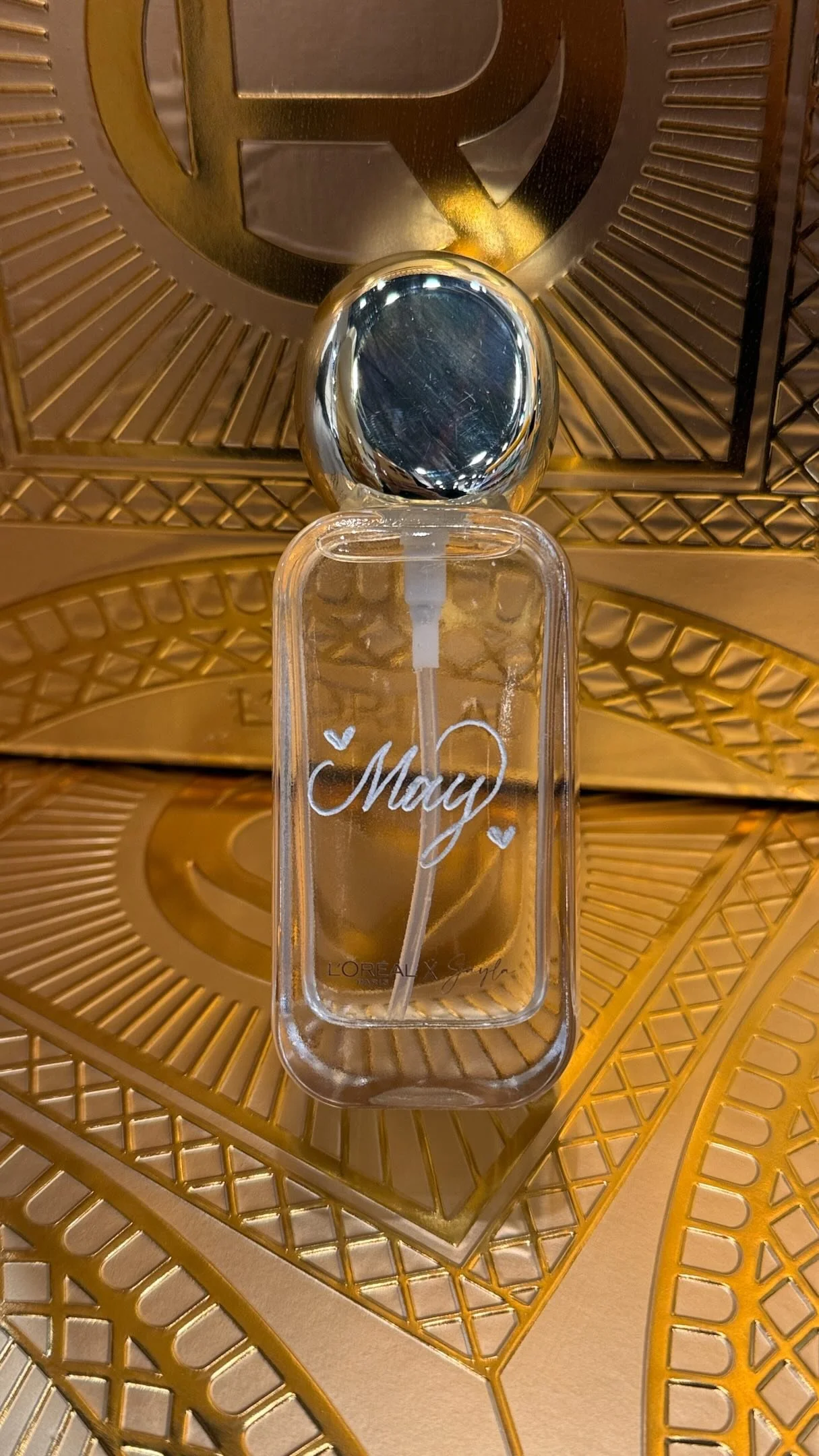 A clear plastic hand sanitizer bottle with the name 'May' written on it in white, resting on a golden decorative surface.