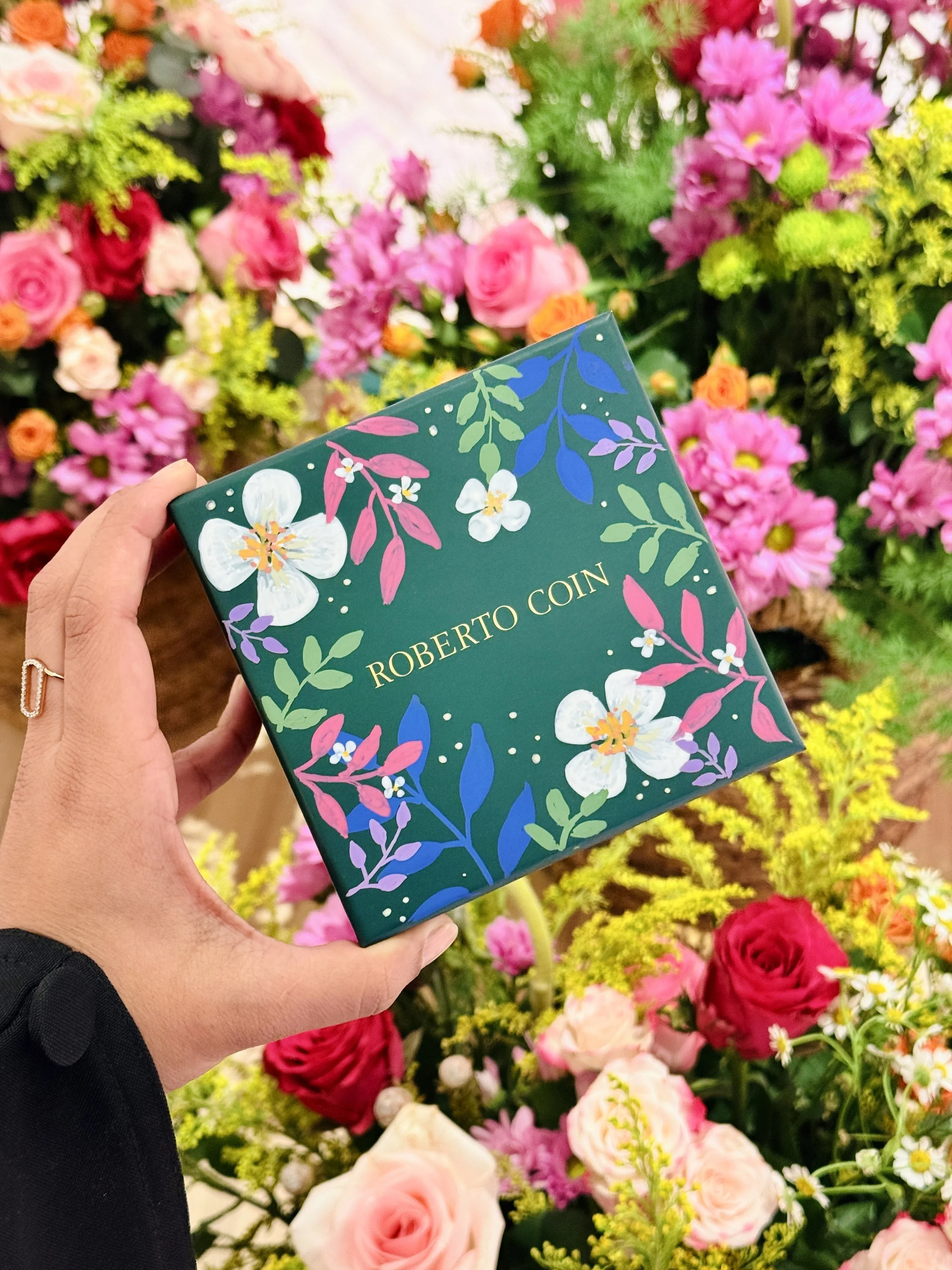 A person holding a floral-patterned box with the word 'Roberto Coin' in front of a colorful display of pink and red flowers.