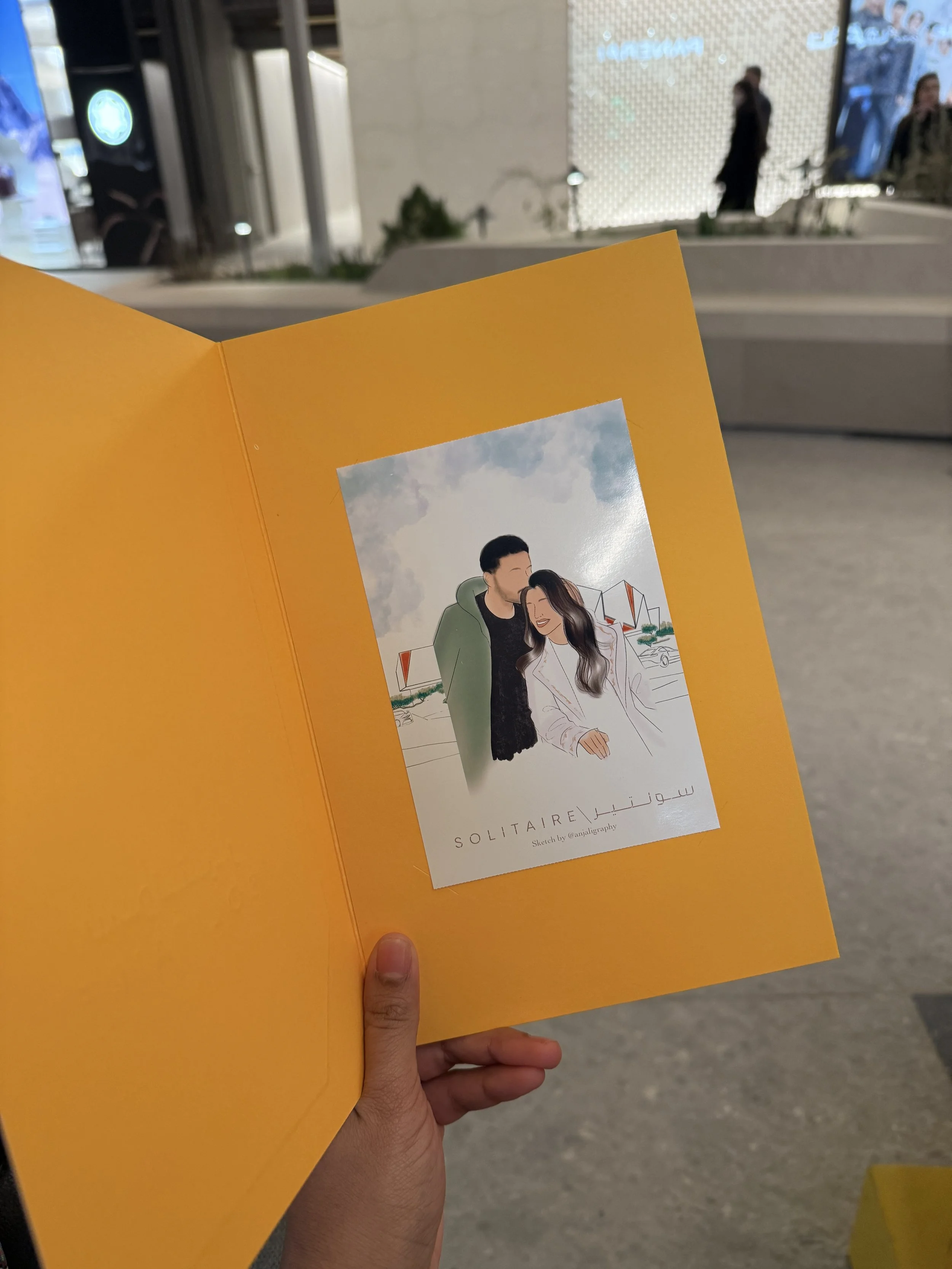 A person holding a yellow brochure with an illustration of a couple on it, in an indoor shopping center or mall.
