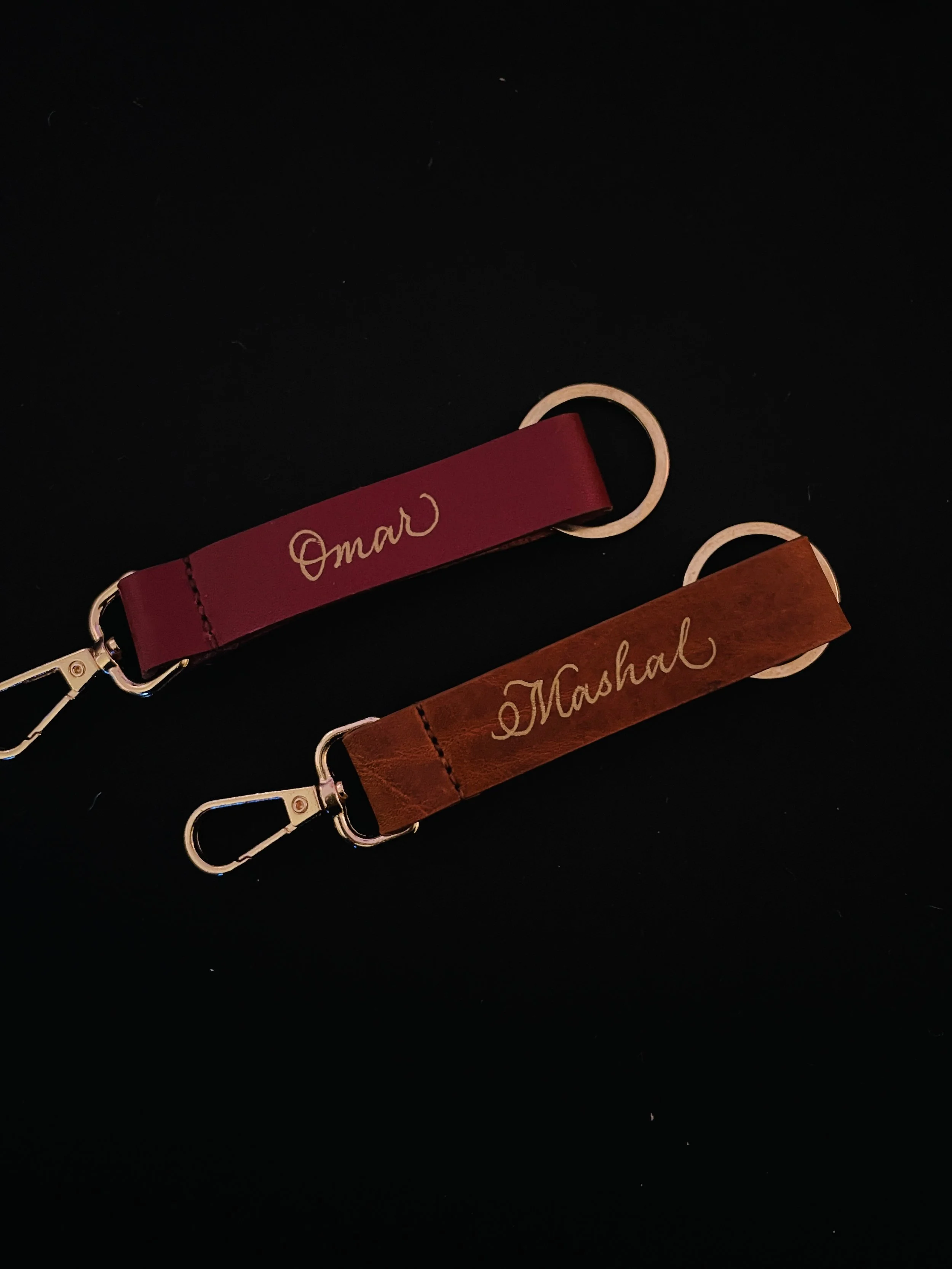 Two leather keychains with metal clips, one maroon with the name 'Omar' and one brown with the name 'Marshal' engraved, on a black background.