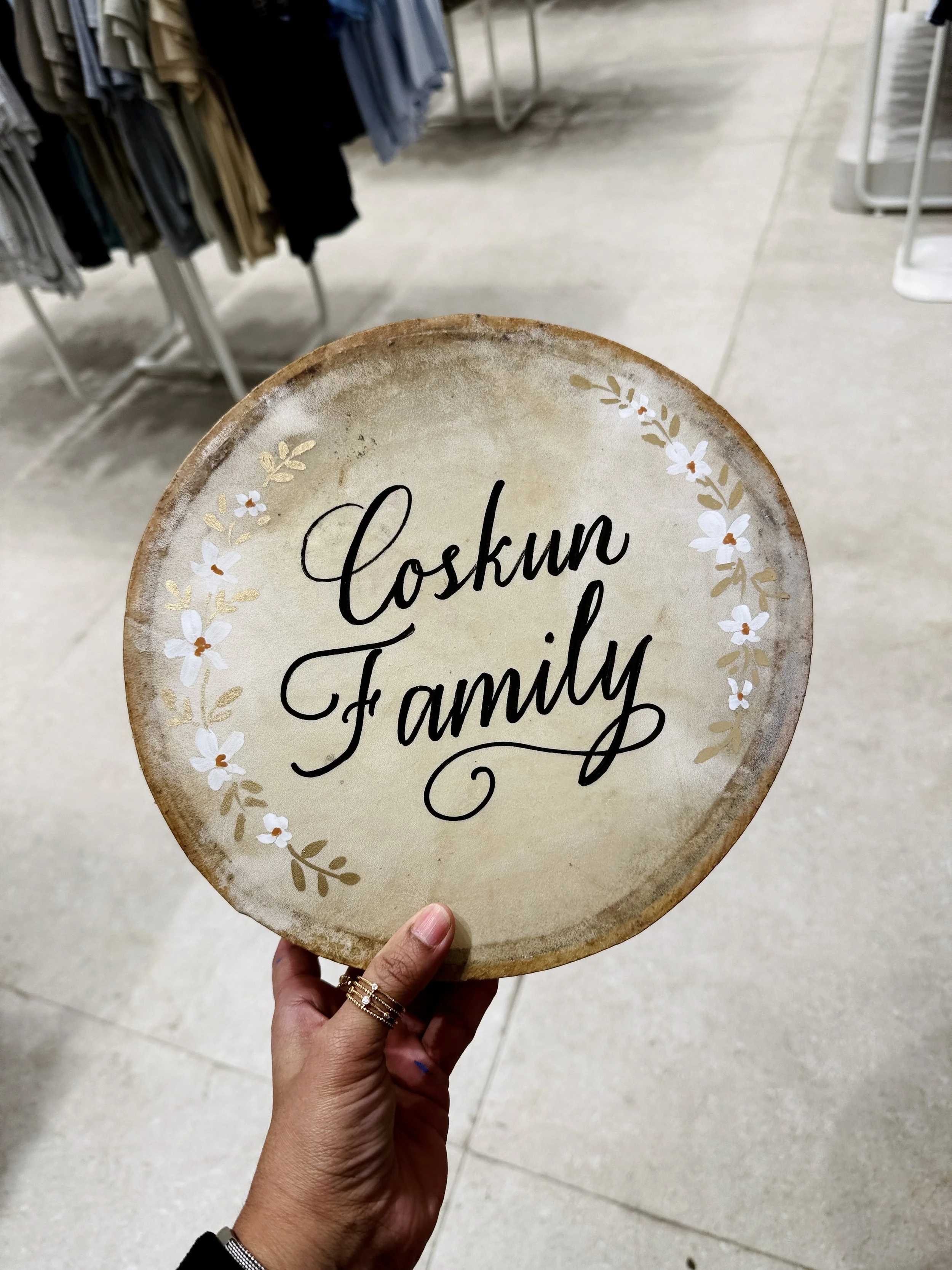 A person holding a decorative round sign that says 'Close for Family' with floral accents around the border. The hand shows multiple rings and a black long-sleeve garment. The background features clothing racks and store tiles.