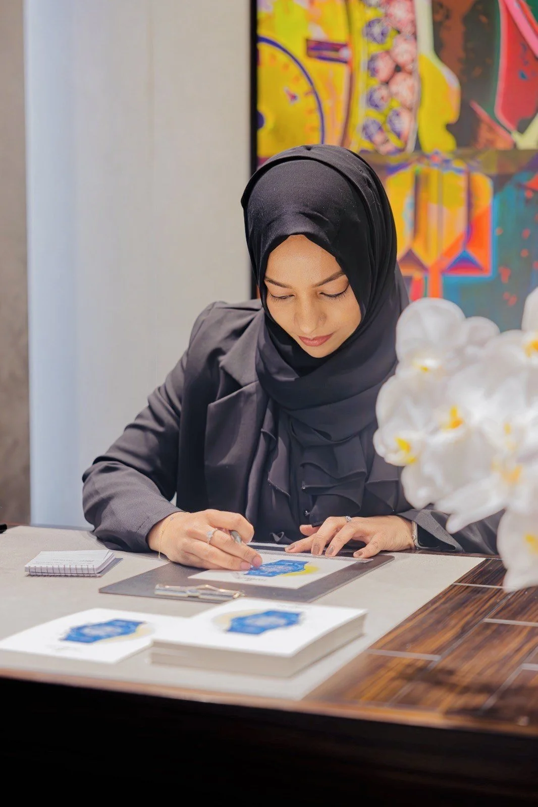 A woman in a dark hijab and gray blazer is sitting at a table, signing or examining artwork. There are art pieces on the table and colorful abstract paintings in the background.