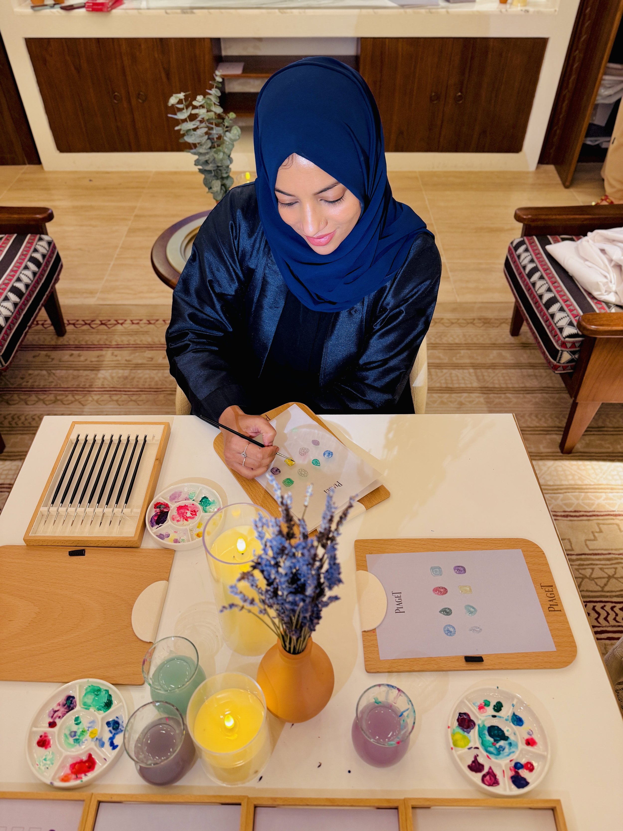 A woman in a blue headscarf and black shirt sitting at a table, painting small stones with colorful paints. The table has palettes with various colors, cups of water, and a yellow vase with purple and yellow flowers. There are chairs with patterned c