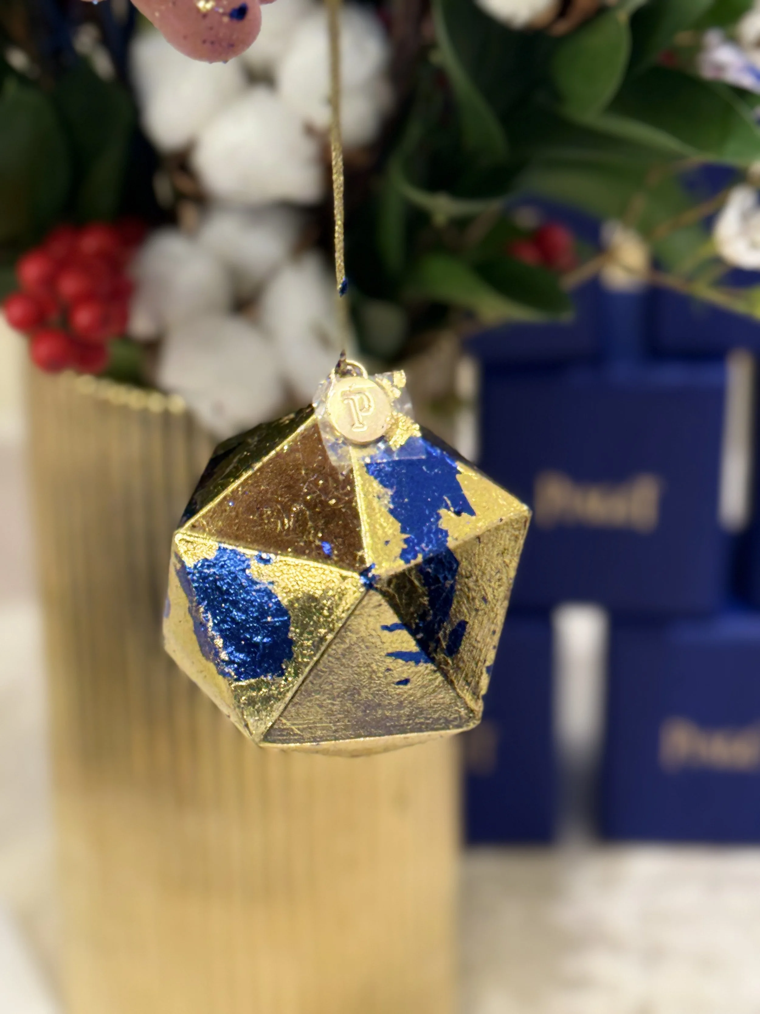 A close-up of a gold and blue decorative hanging ornament with a small circular tag marked with a 'P', hanging in front of holiday greenery and white berries.