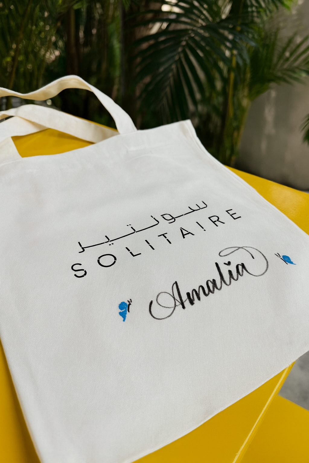 White tote bag with Arabic and English text: 'Sooltairé' and 'Amalia', placed on a yellow surface with green plants in the background.
