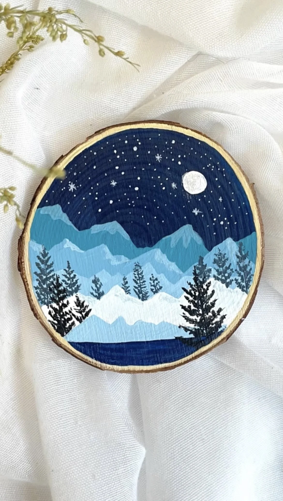Decorative wooden slice with a nighttime mountain landscape scene painted on it, featuring snow-covered mountains, pine trees, a starry sky, and a full moon, resting on white fabric with a sprig of small yellow flowers nearby.