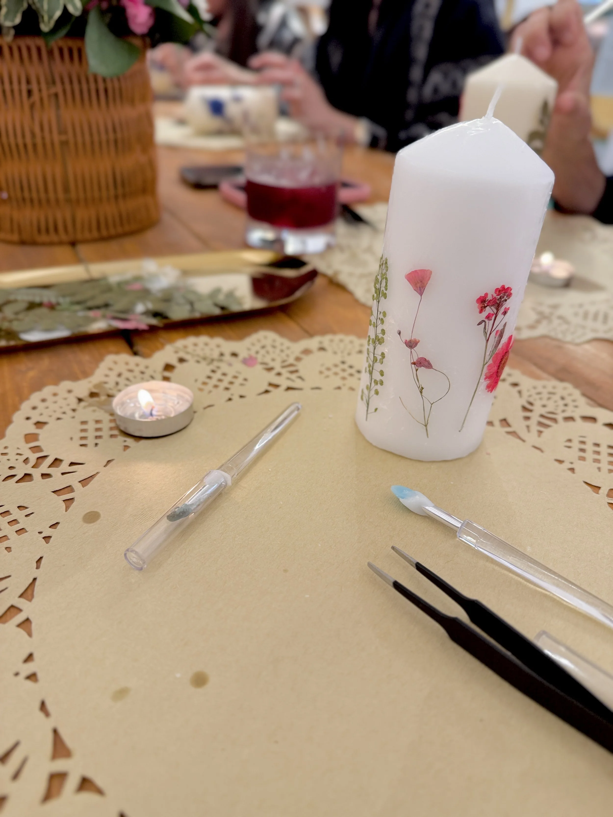 A white candle with a floral design on it, placed on a yellow doily, with a small tealight candle nearby. There are dental tools resting on the doily, and a wooden table with a tray, drinks, and a person sitting in the background.
