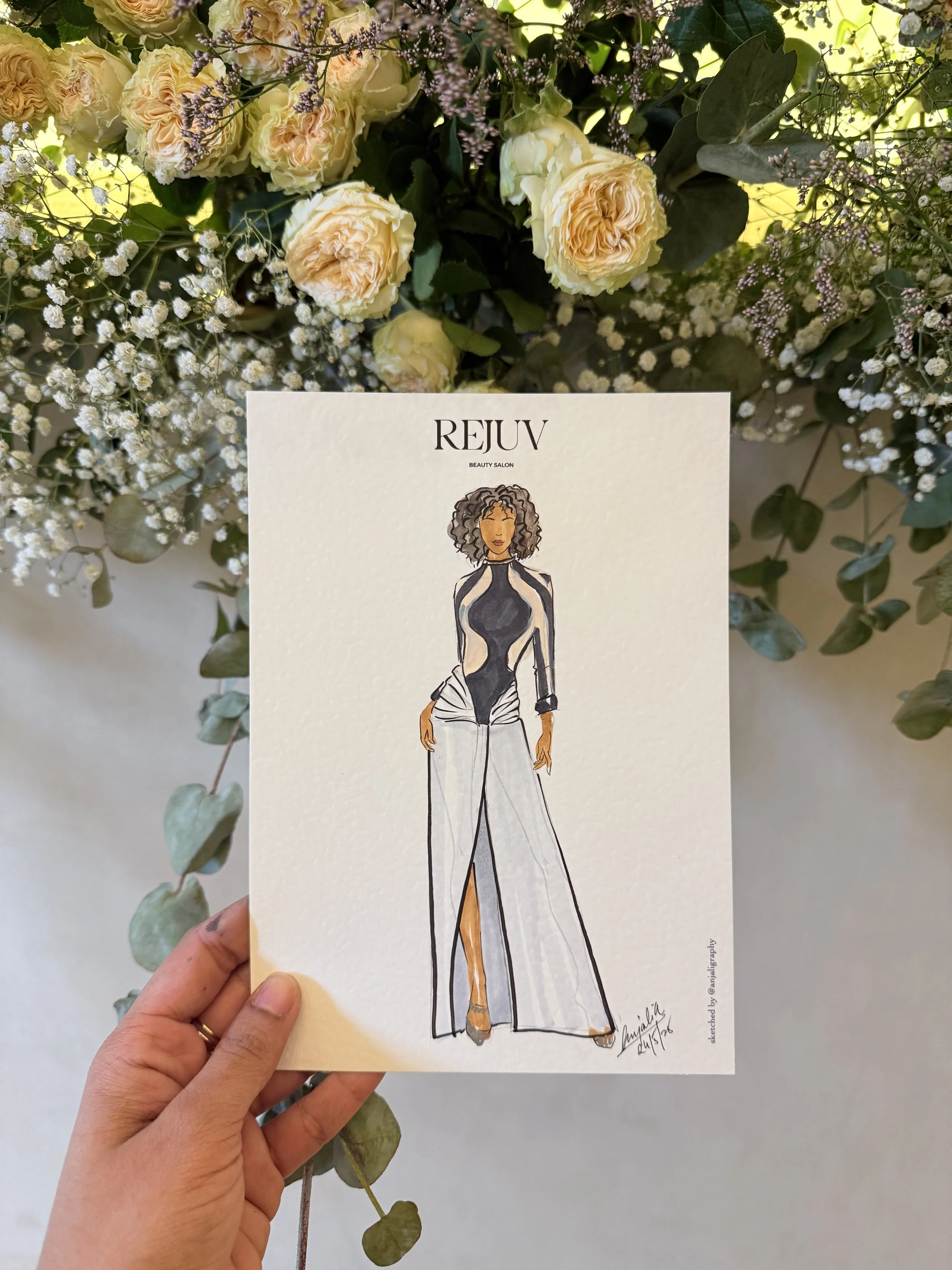 A hand holding a fashion illustration card featuring a woman in a black and white outfit, in front of a floral arrangement with cream roses, eucalyptus leaves, and small purple and white flowers.