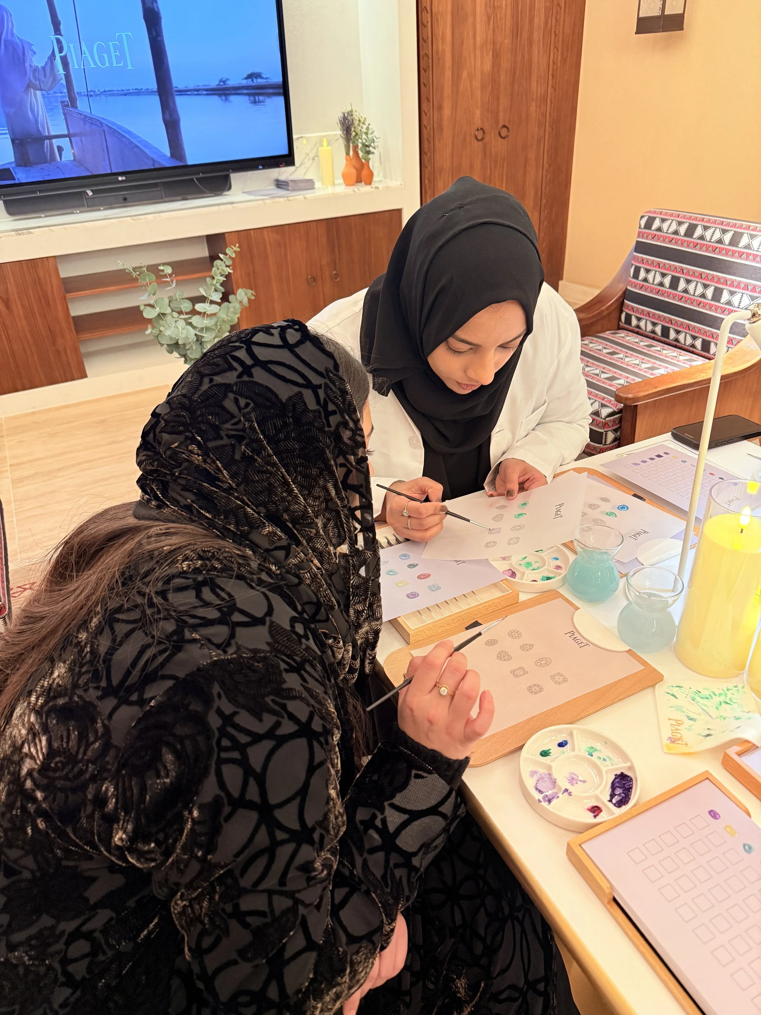 Two women wearing headscarves painting designs on paper with small brushes, surrounded by paint palettes and other arts and crafts materials in a cozy room.