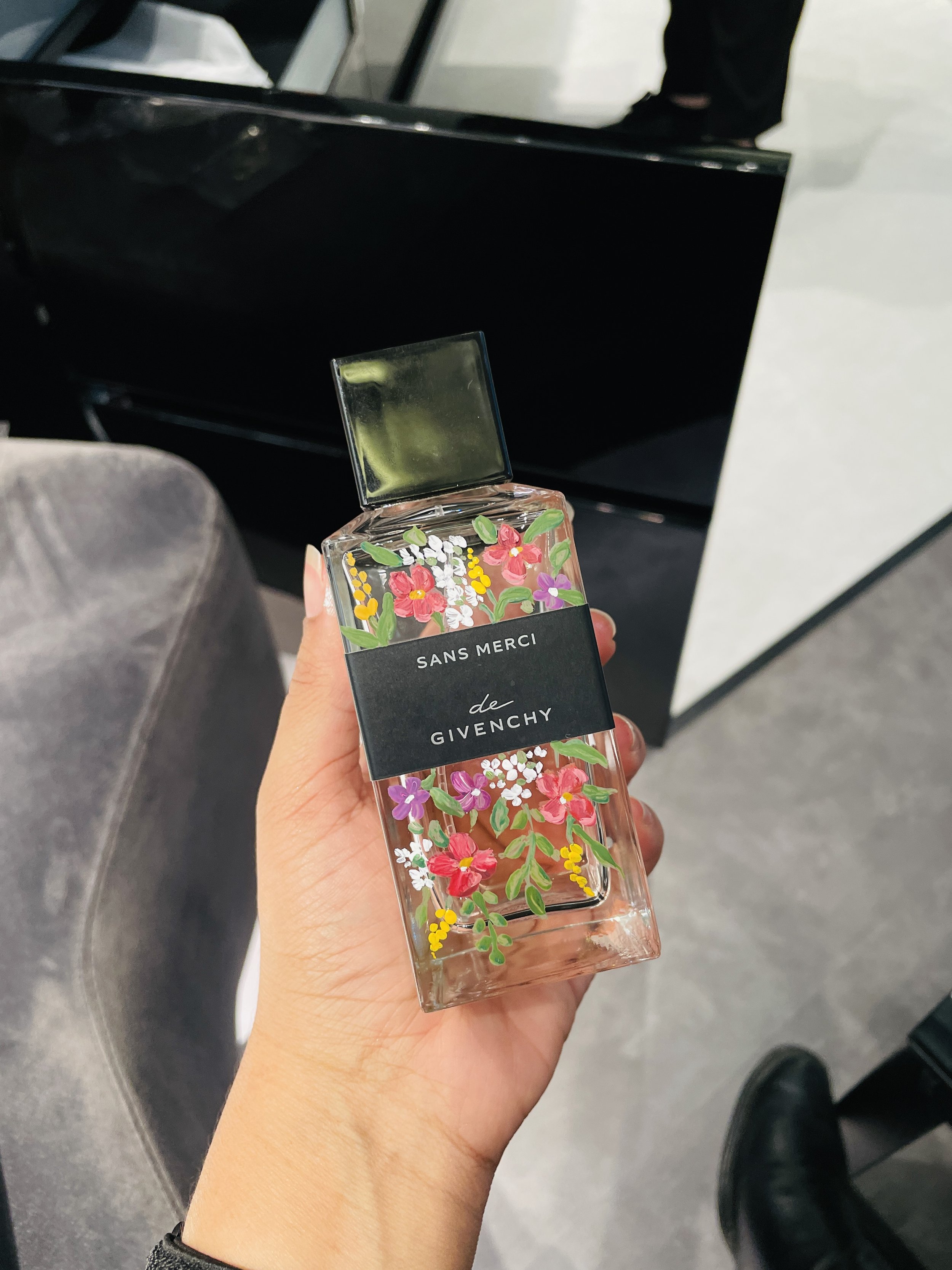 Hand holding a floral-themed perfume bottle labeled 'SANS MERCI de GIVENCHY' with a black cap, in a store with gray floor and a black display or counter in the background.