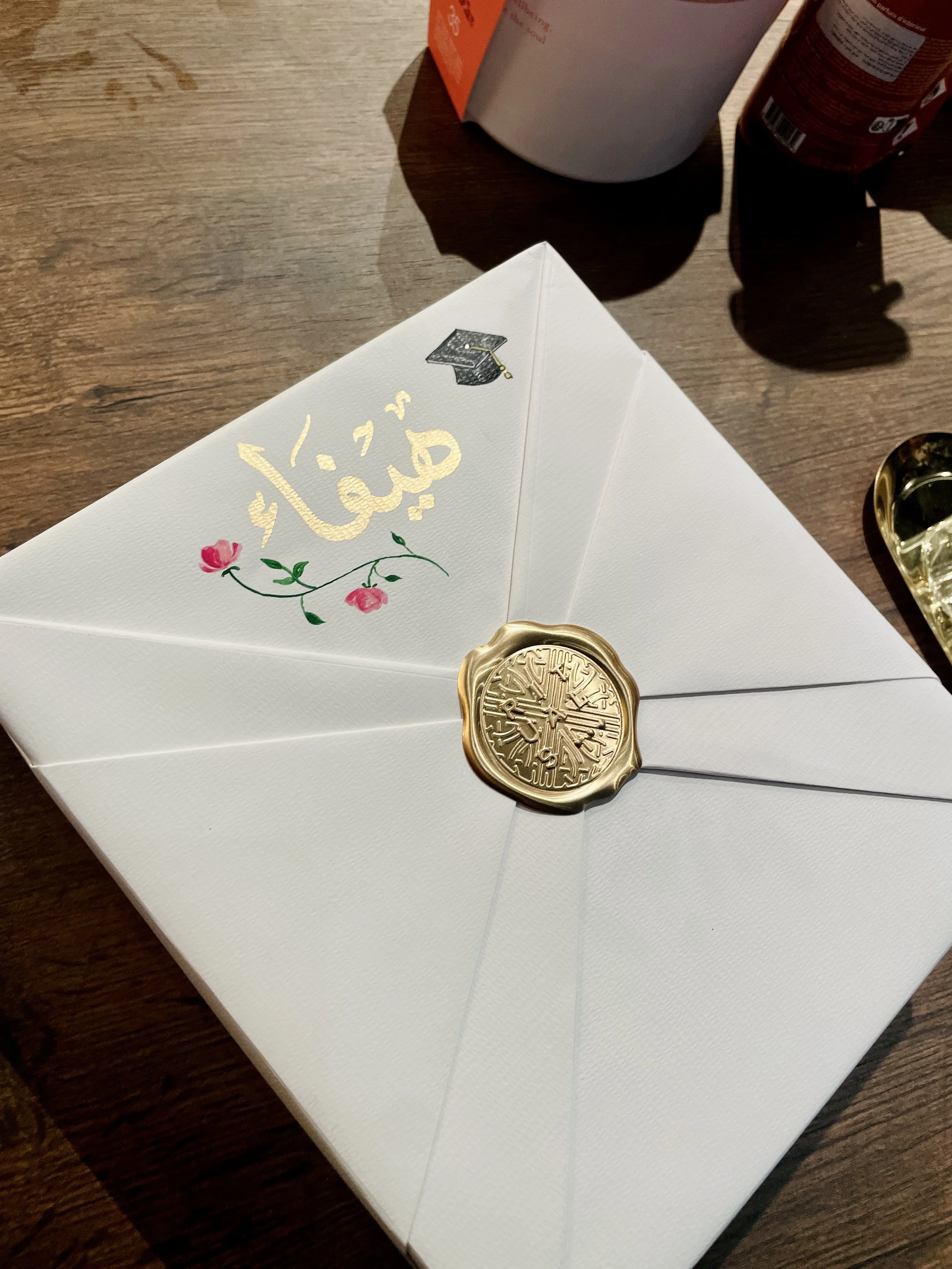 An envelope decorated with a pink flower and gold writing, sealed with a gold wax seal, resting on a wooden surface alongside jars and containers.