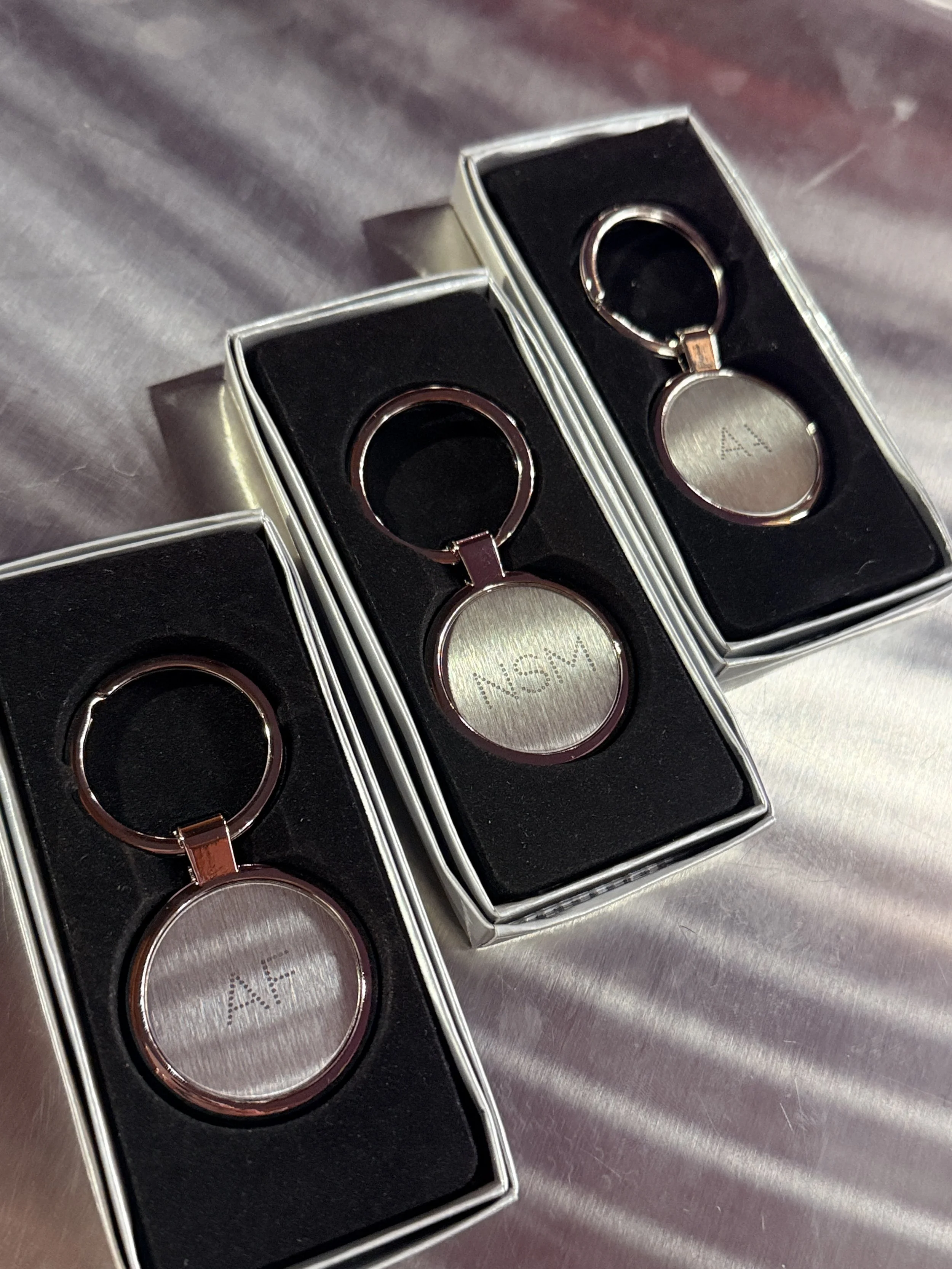 Two metallic keychains with circular tags, one with 'AF' engraved, placed in black velvet-lined boxes on a metallic surface.
