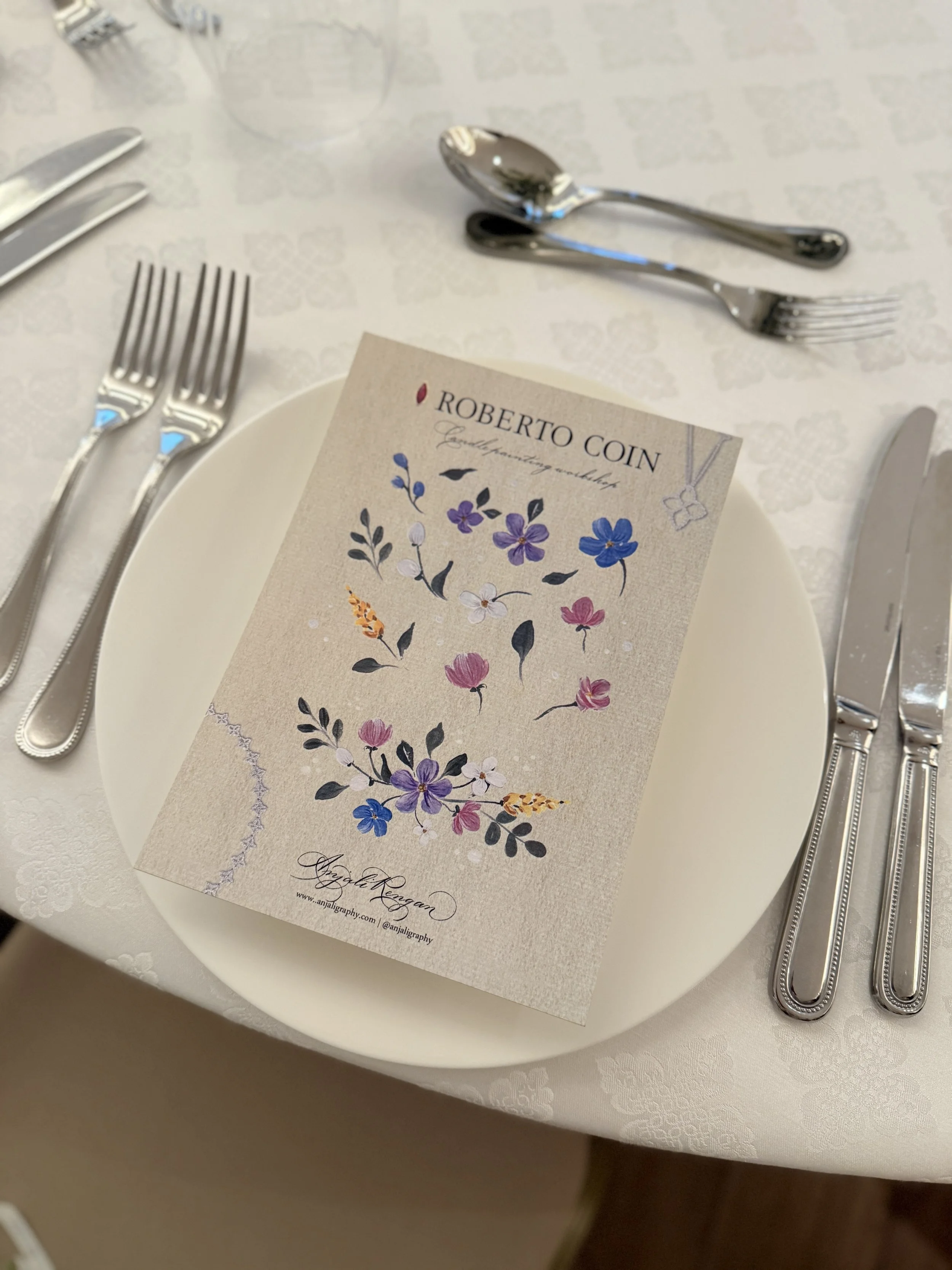 A table setting with a white plate, three forks on the left, three knives and spoons on the right, a napkin with a floral design, and a card with the name 'Roberto Coin' on a white tablecloth.