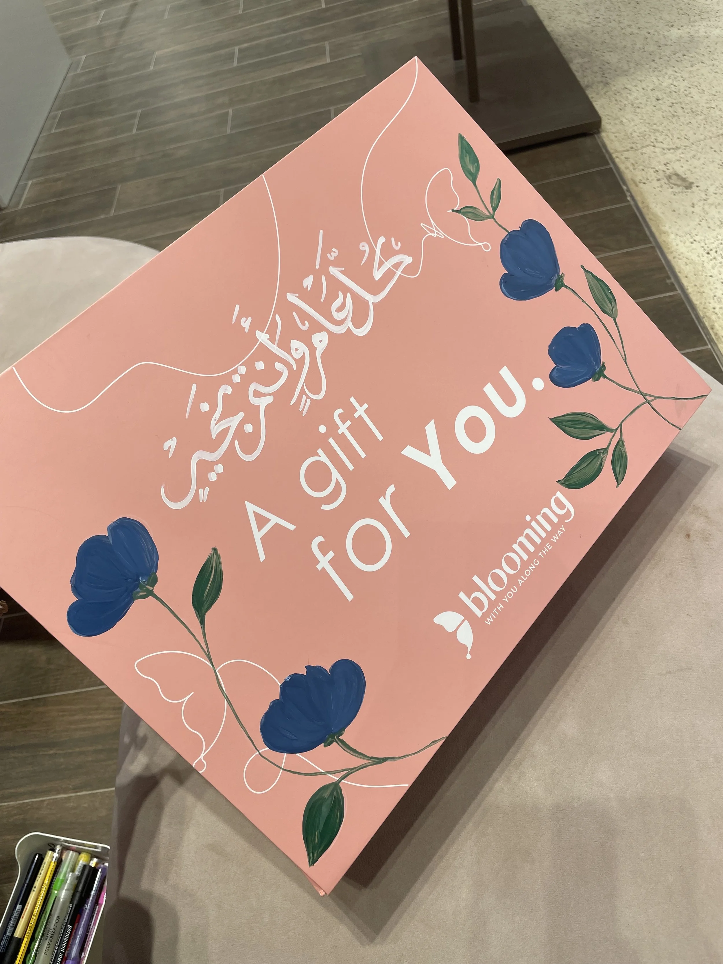 A pink poster with white Arabic calligraphy and blue and green floral designs. The poster has text in English that reads: "A gift for you from Bloom Way".