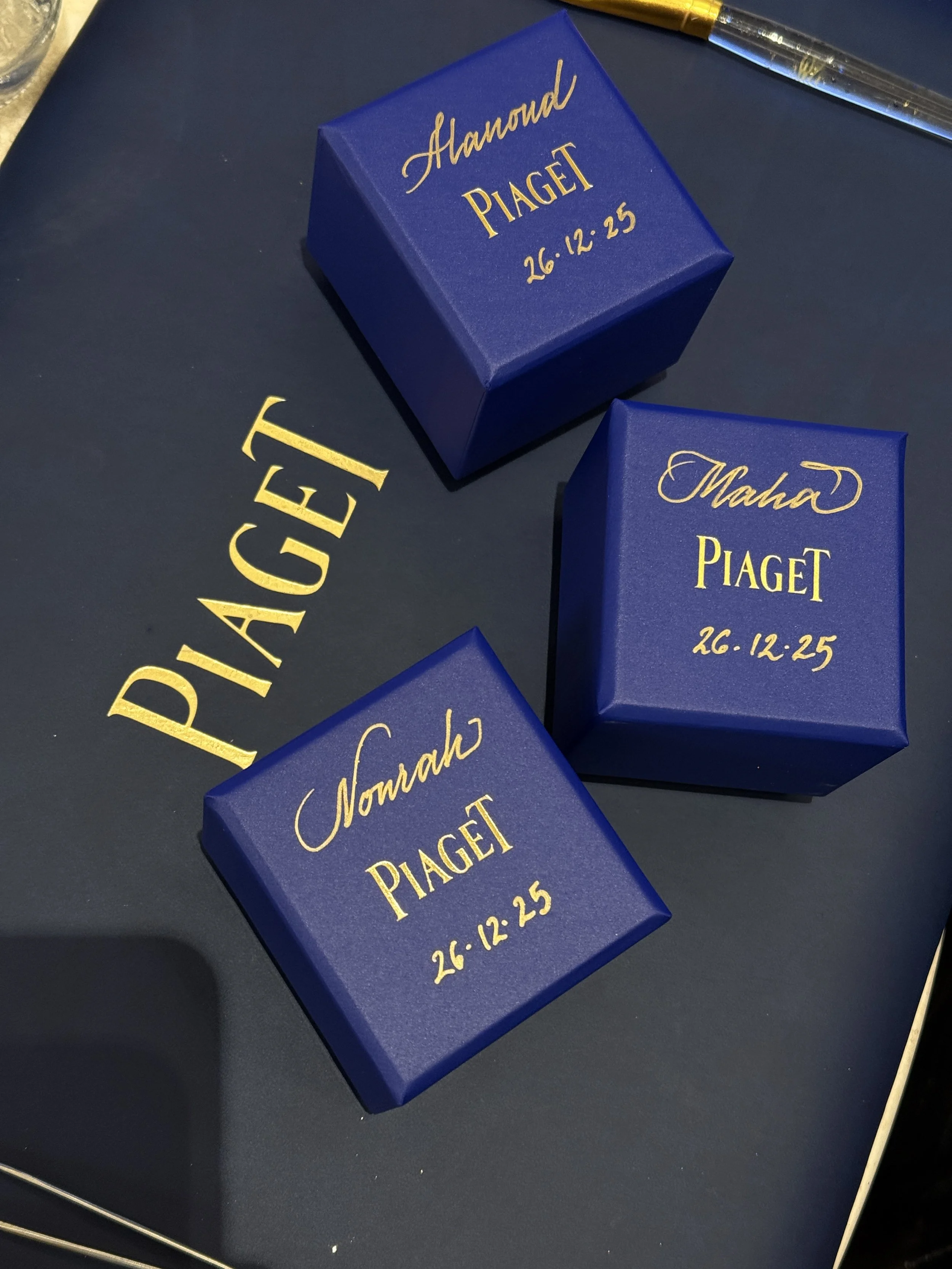 Blue boxes with gold text reading 'Honour Piaget' and a date '26.12.25' on a navy blue surface.