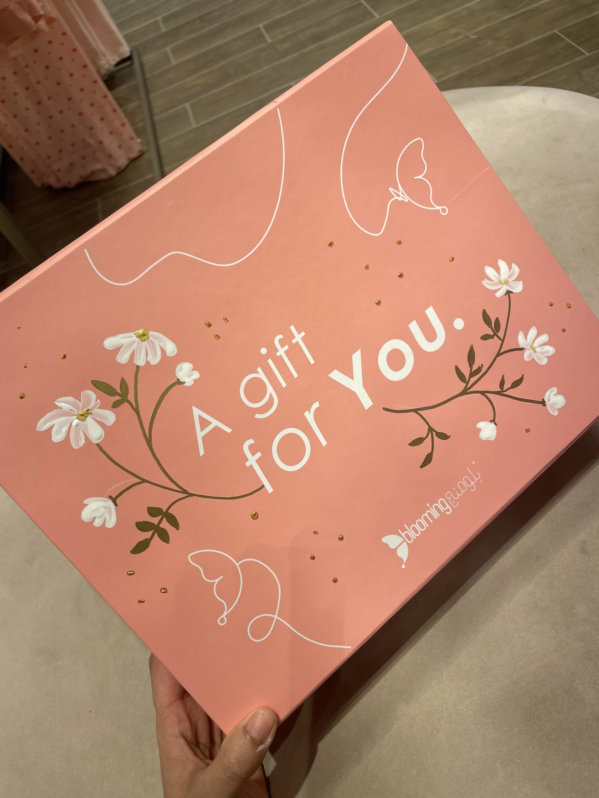 Pink gift box with white floral illustrations and text, held in a person's hand.