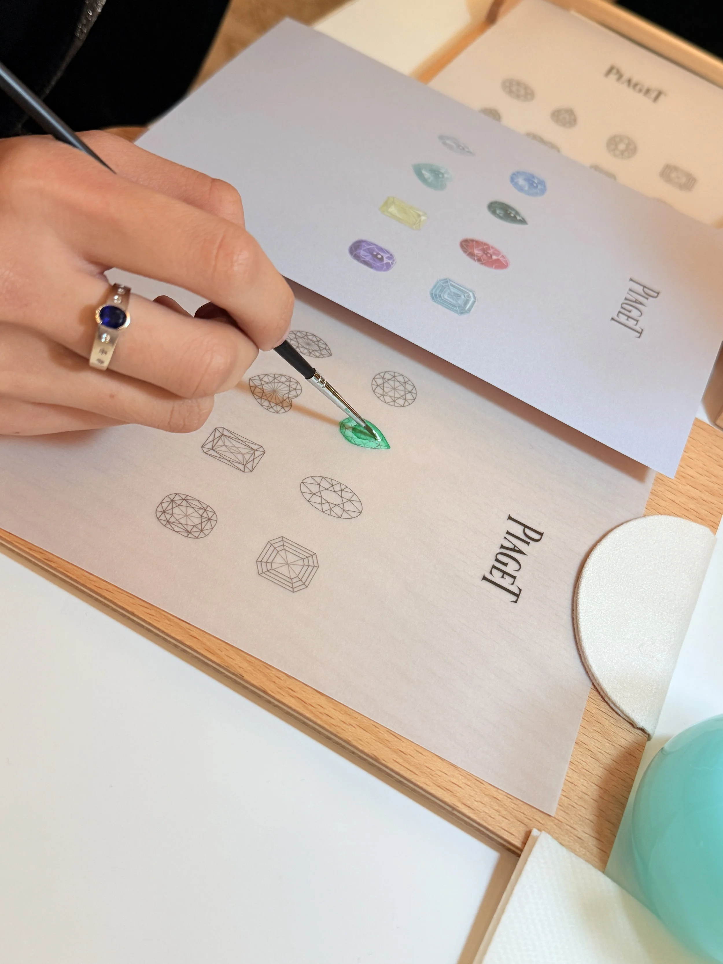 Close-up of a person's hand holding a fine-tip brush and painting a green gemstone illustration on a piece of paper. A ring with a blue gemstone is on their finger. There are various gemstone illustrations and a booklet labeled 'PAGET' in the backgro