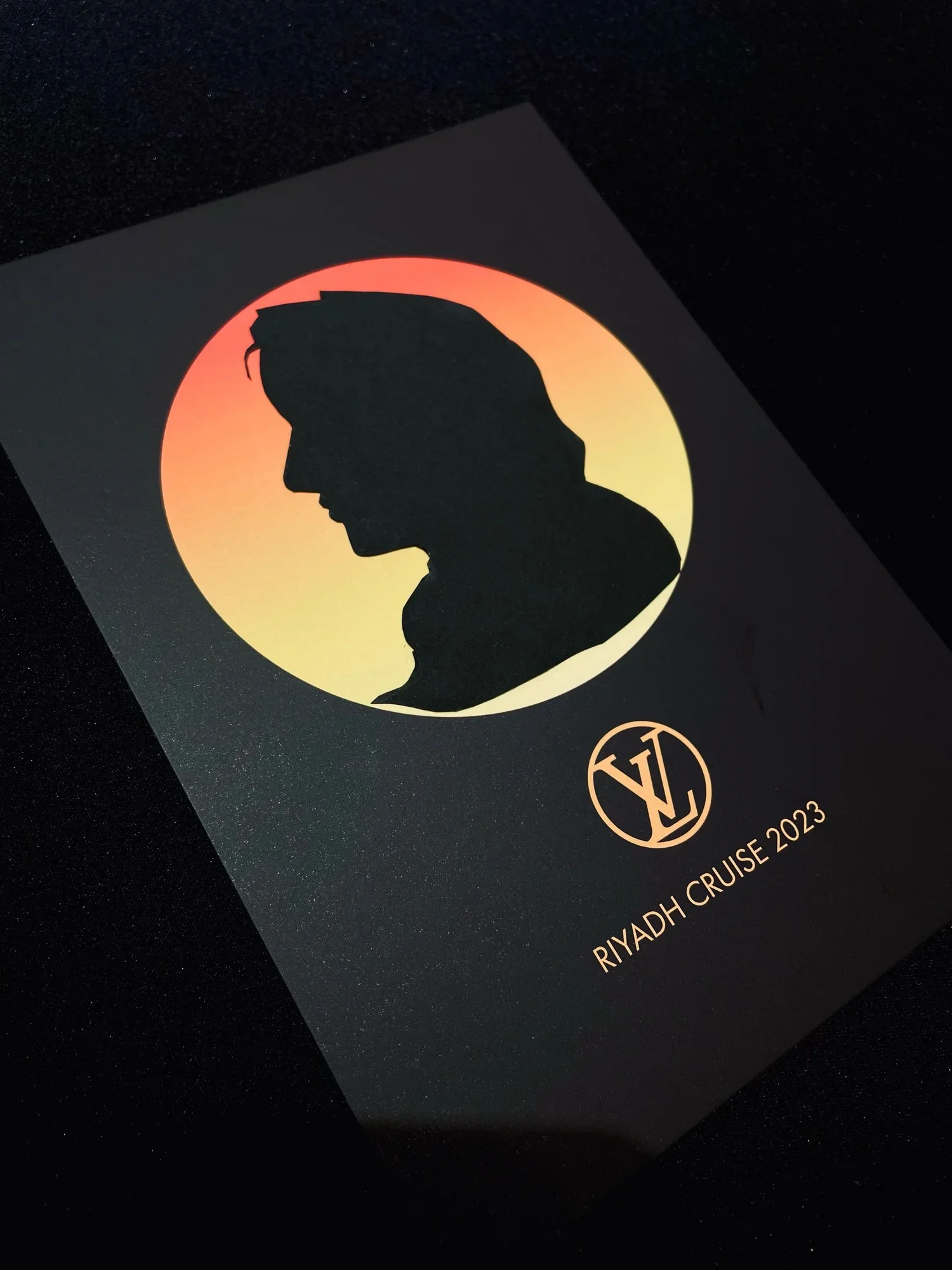 A black card features a silhouette of a person's profile with a sunset or sunrise background in gradient hues of red, orange, and yellow. The card includes the text "RIYADH CRUISE 2023" and a circular logo with the initials "V" and "K".