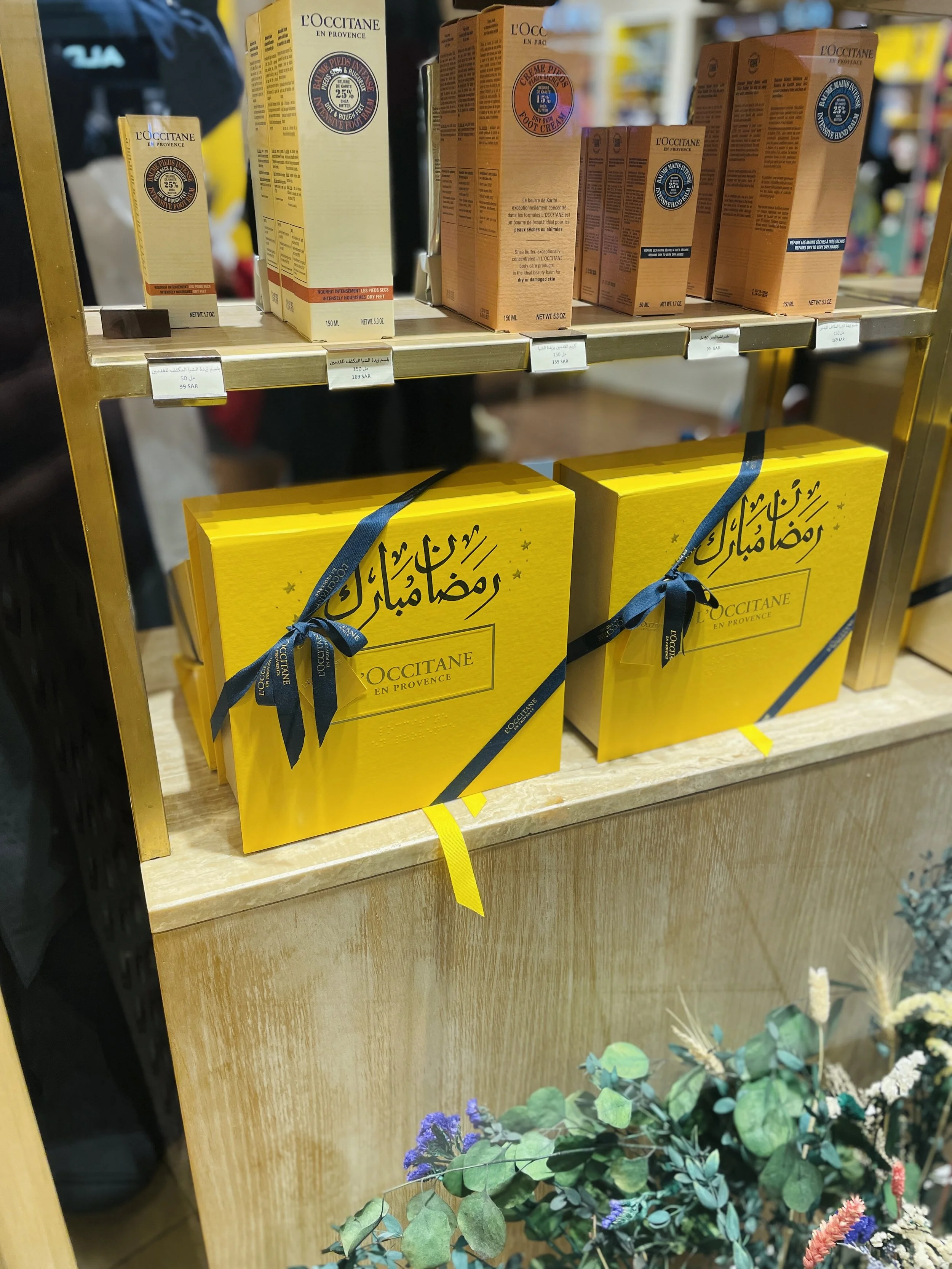 Various L'Occitane skincare products displayed on a wooden shelf, with two yellow gift boxes with Arabic writing and black ribbons in front, and decorative flowers in the foreground.