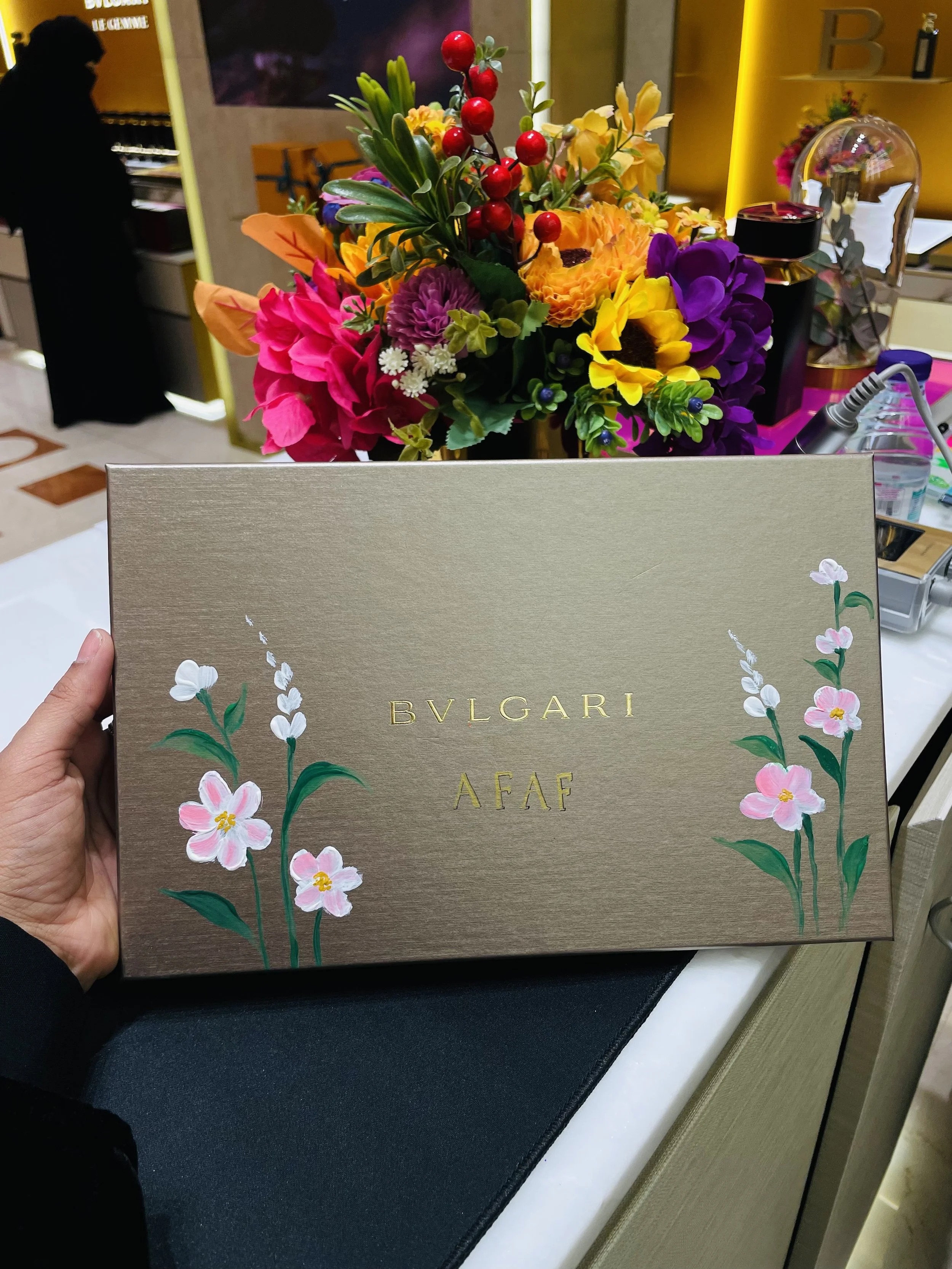 A hand holding a beige book with floral design and gold "BVLGARI AFAF" text in front of a colorful flower arrangement on a table.