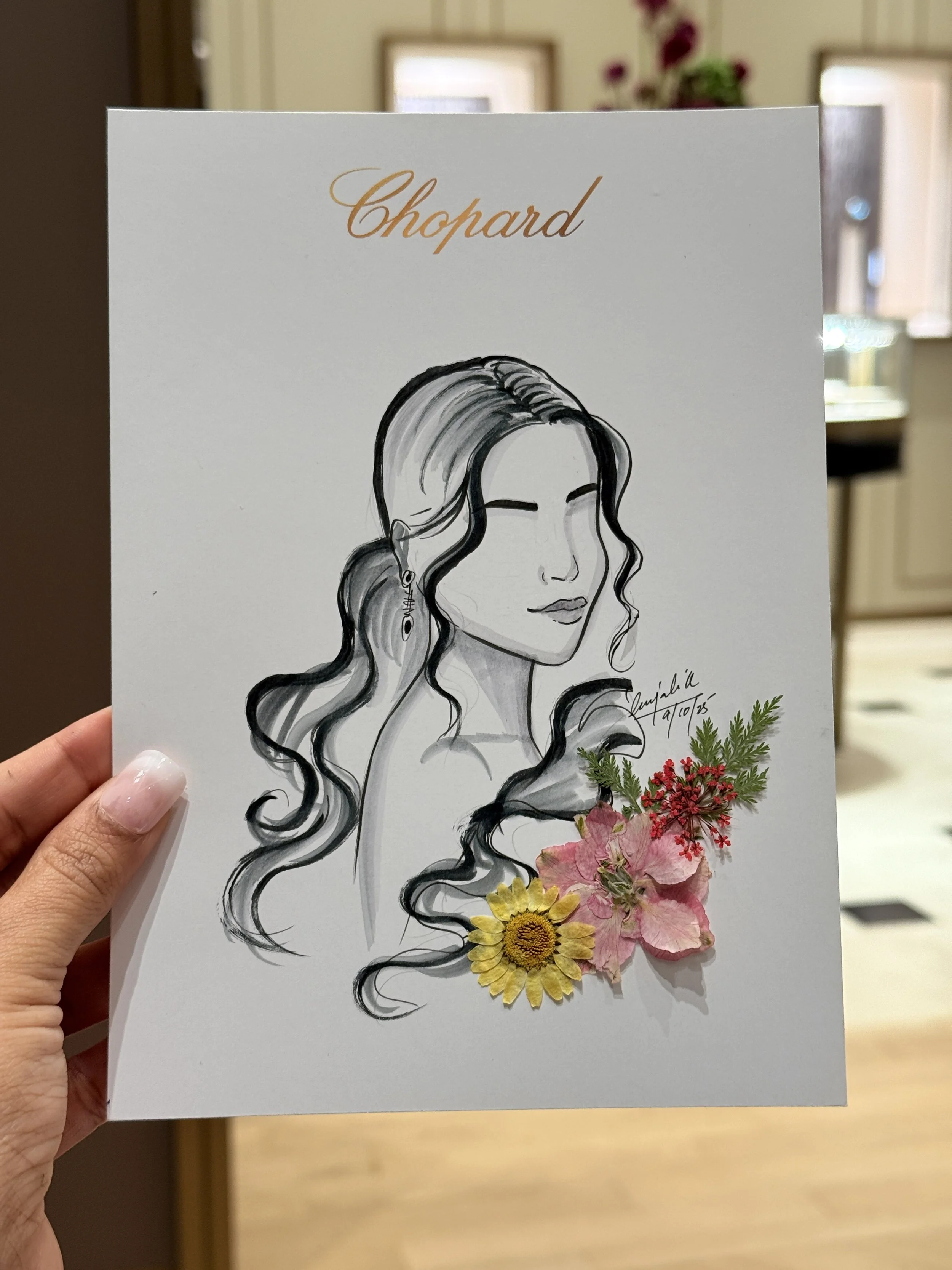 Hand holding a white card with a black and white line drawing of a woman with long wavy hair and closed eyes, decorated with pressed flowers at the bottom, with the word 'Chopard' written in cursive at the top.