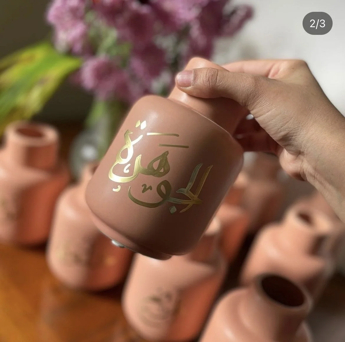 A hand holding a pink candle with gold Arabic calligraphy, with several similar candles in the background and pink flowers in the blurred background.