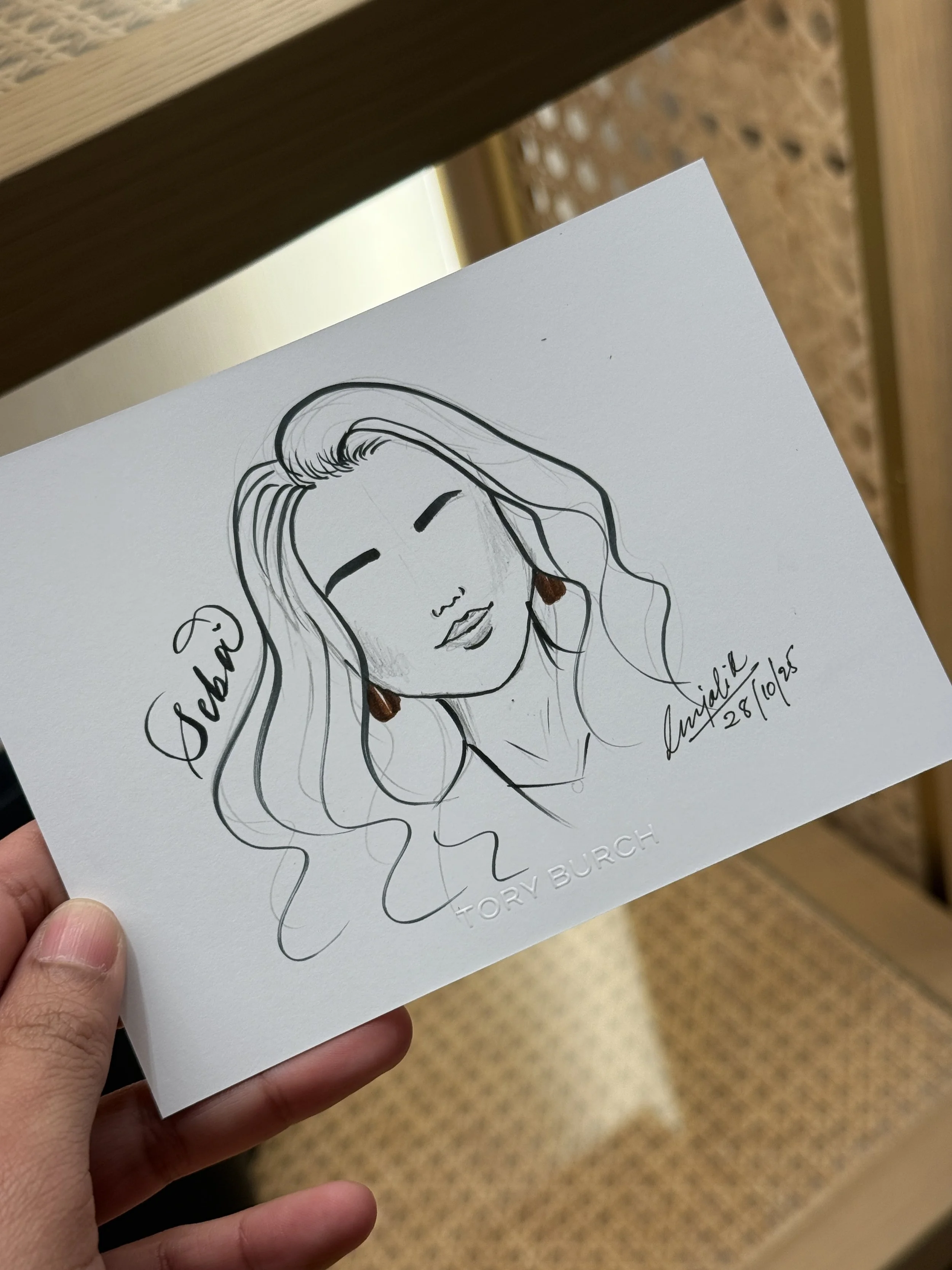 A hand holding a white paper featuring a line drawing of a woman's face with long hair, earrings, and closed eyes. The drawing is signed by Tory Burch and dated October 28, 2016.
