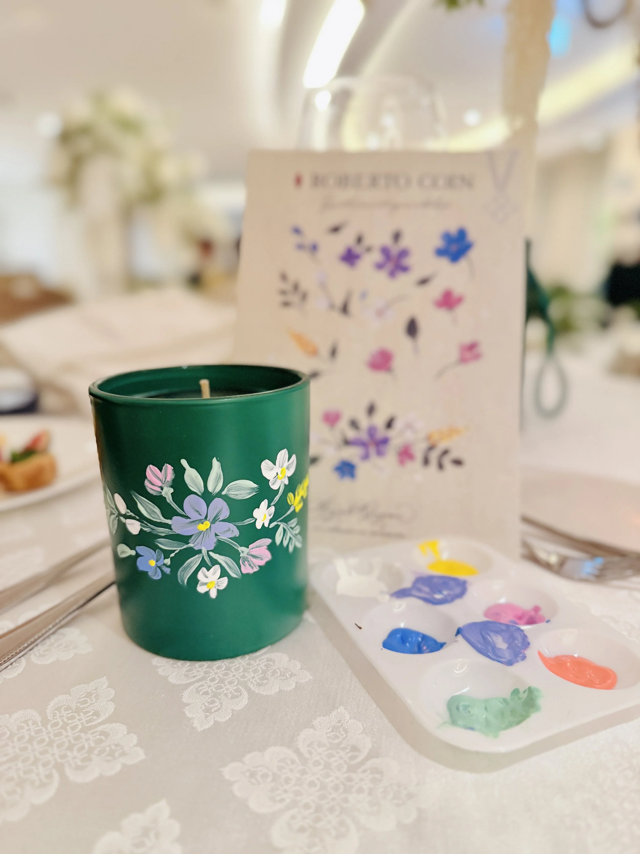 A teal green candle holder decorated with colorful flowers and white leaves. Next to it is a white card with watercolor flowers. The table has paint colors and brushes, suggesting a painting or craft activity.