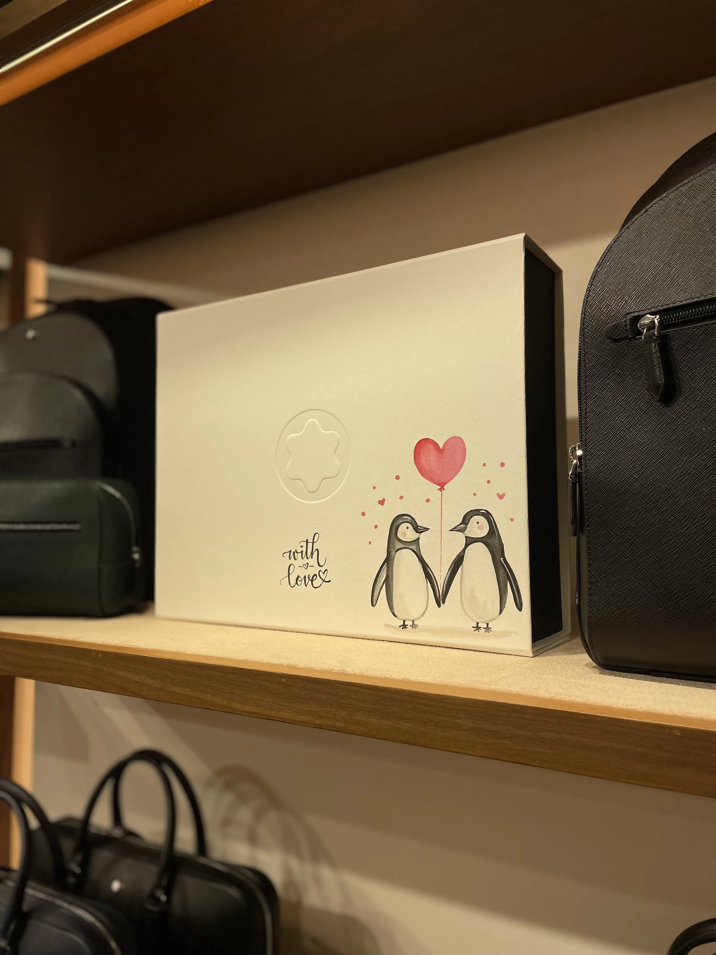Shelf with a greeting card featuring two cartoon penguins holding a heart-shaped balloon and the words "with love." Black backpacks and a brown bookcase are also visible.