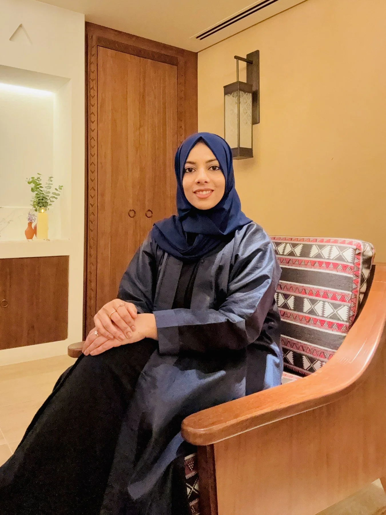 A woman wearing a blue hijab and gray jacket sitting at a wooden table in a warmly lit room, smiling at the camera.