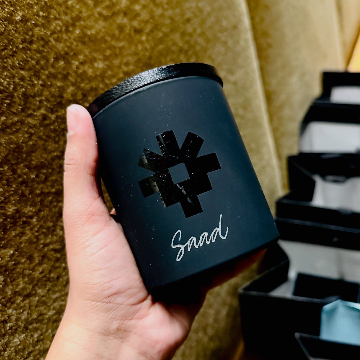 Hand holding black container labeled 'Snad' with geometric black logo, against a textured brown background.
