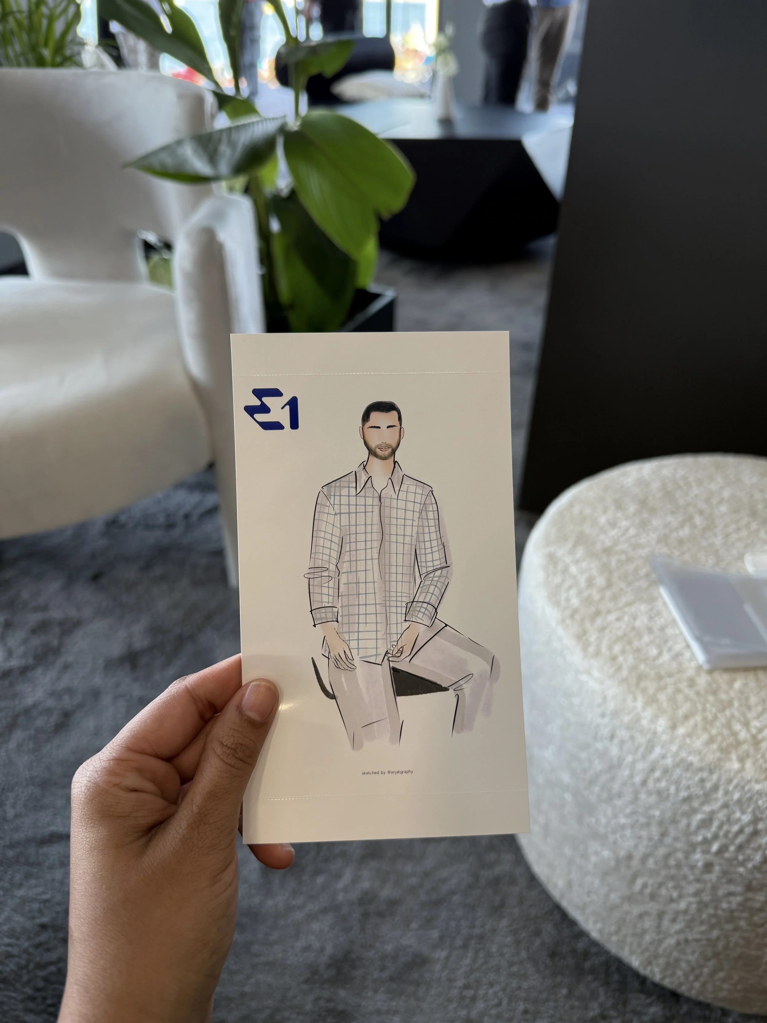 A hand holding a piece of paper with a stylized drawing of a man sitting on a chair at a table. In the background, there is a green plant, a white chair, and some furniture in a room with large windows.