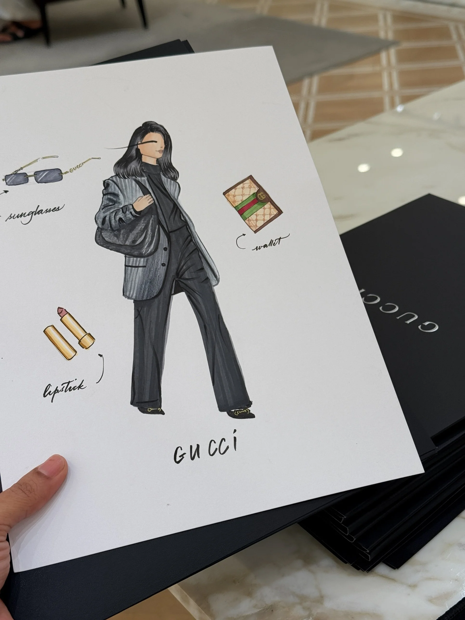 Fashion illustration of a woman in a gray suit with black hair, wearing sunglasses, with a Gucci wallet and lipstick nearby.