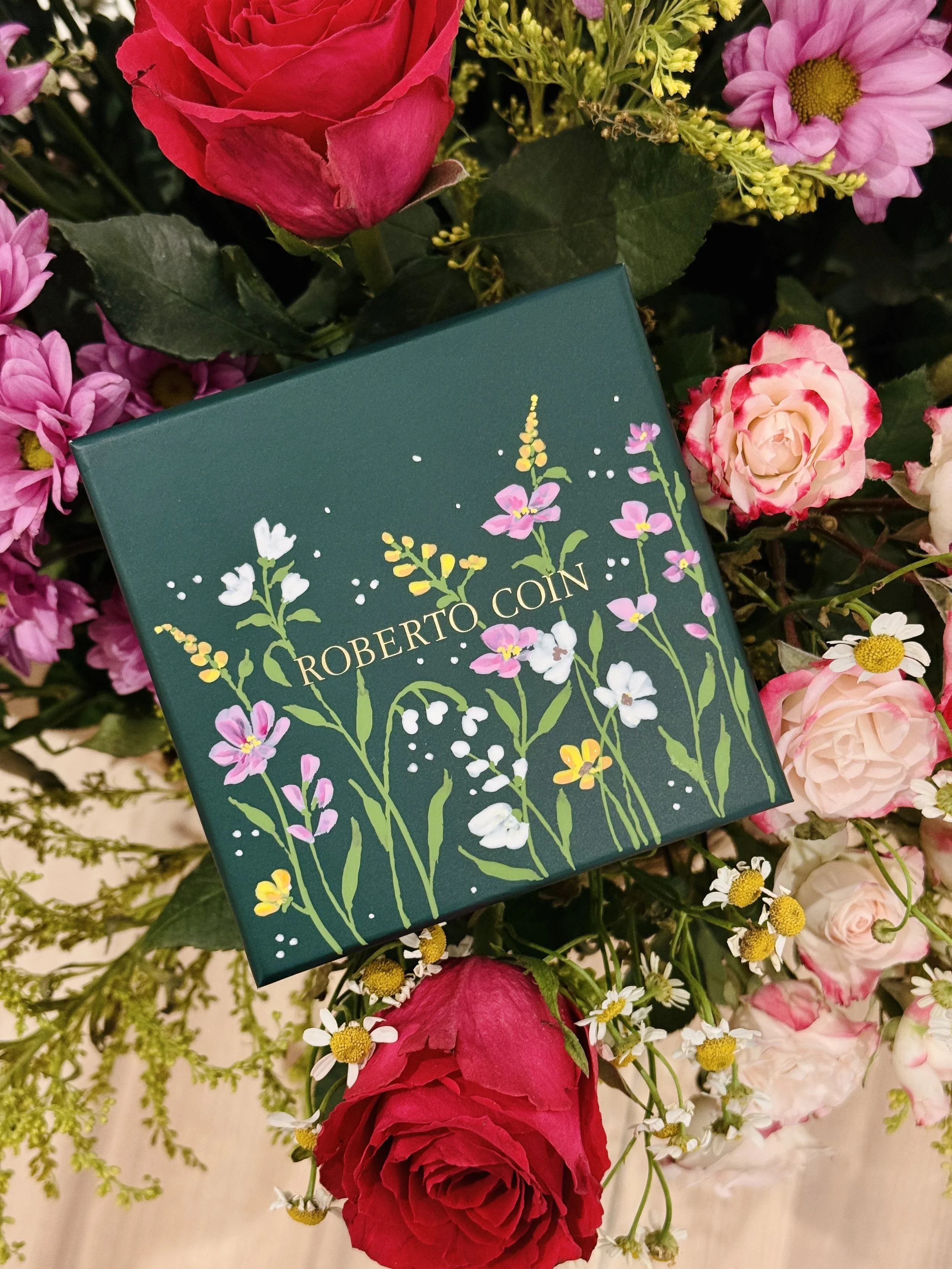 A colorful arrangement of pink, red, and white flowers includes roses and daisies, with a dark green book with floral illustrations placed on top.