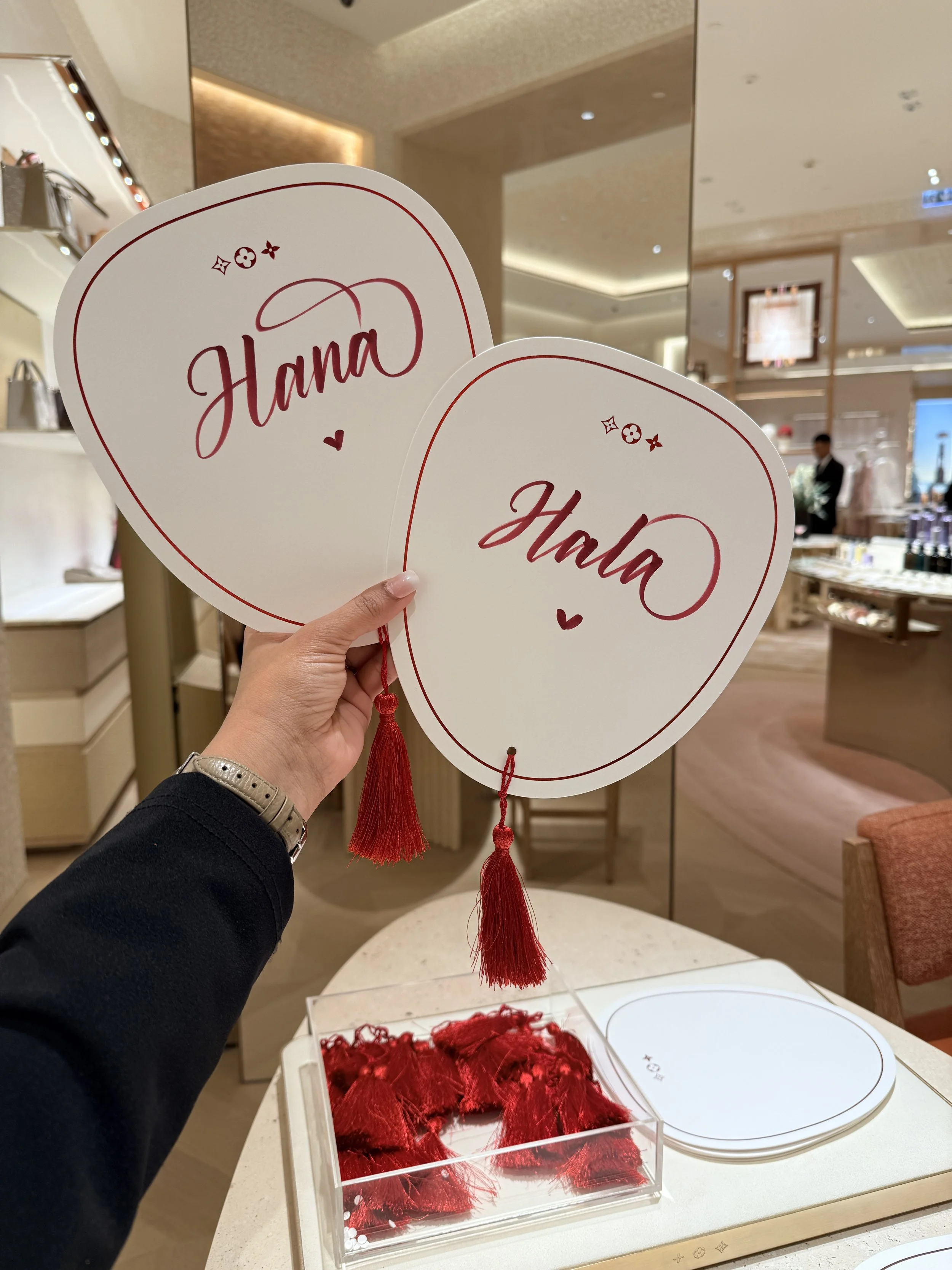 Two white heart-shaped fans with red text 'Hana' and small hearts, held by a person in a store with shelves of handbags and jewelry in the background.