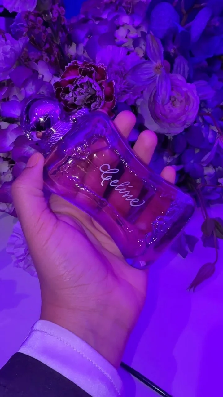 A hand holding a purple-tinted glass bottle with the word "Welcome" written on it, surrounded by purple flowers under purple lighting.