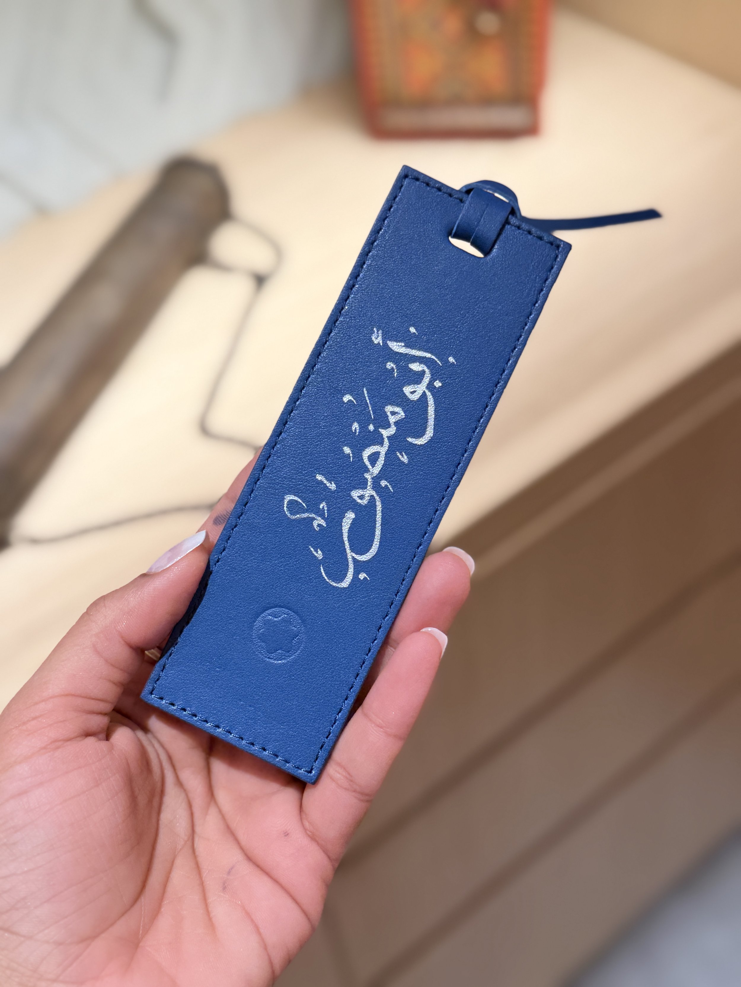 A hand holding a blue leather keychain with Arabic script written in white on it. In the background, there is a blurred table with a rectangular object and a cylindrical item.