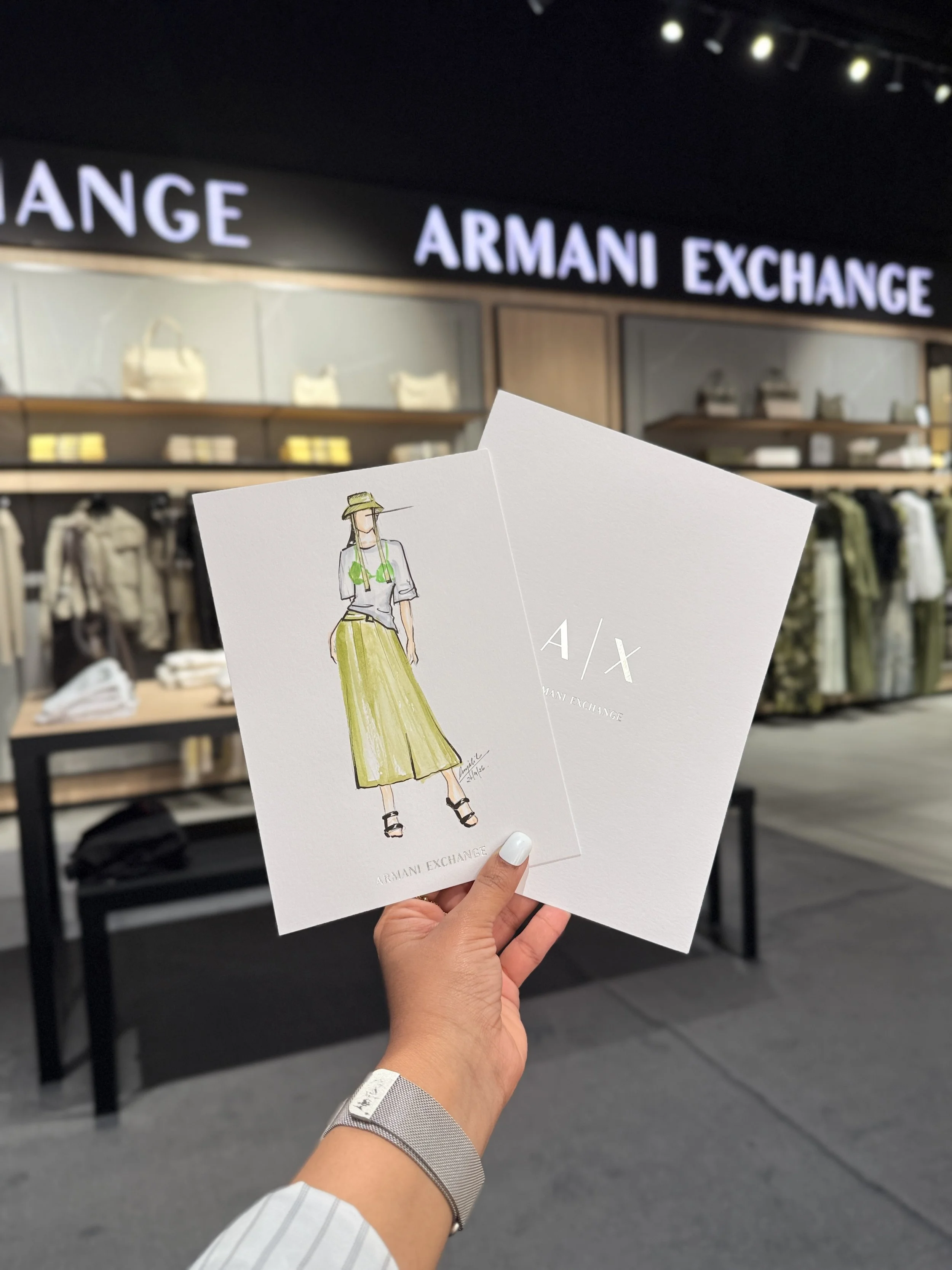 Person holding fashion branding cards in front of Armani Exchange storefront with clothing displays in the background.