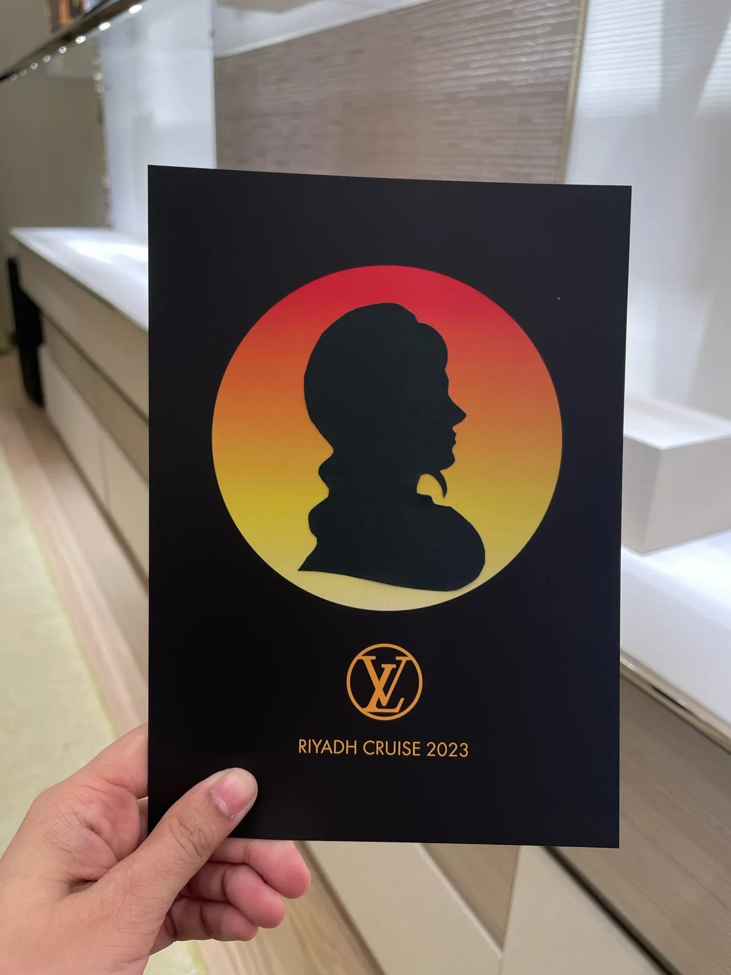 A person holding a black brochure with a silhouette of a woman with a bun and long hair in front of a sunset circle. The text reads "RIYADH CRUISE 2023" and a Louis Vuitton logo at the bottom.