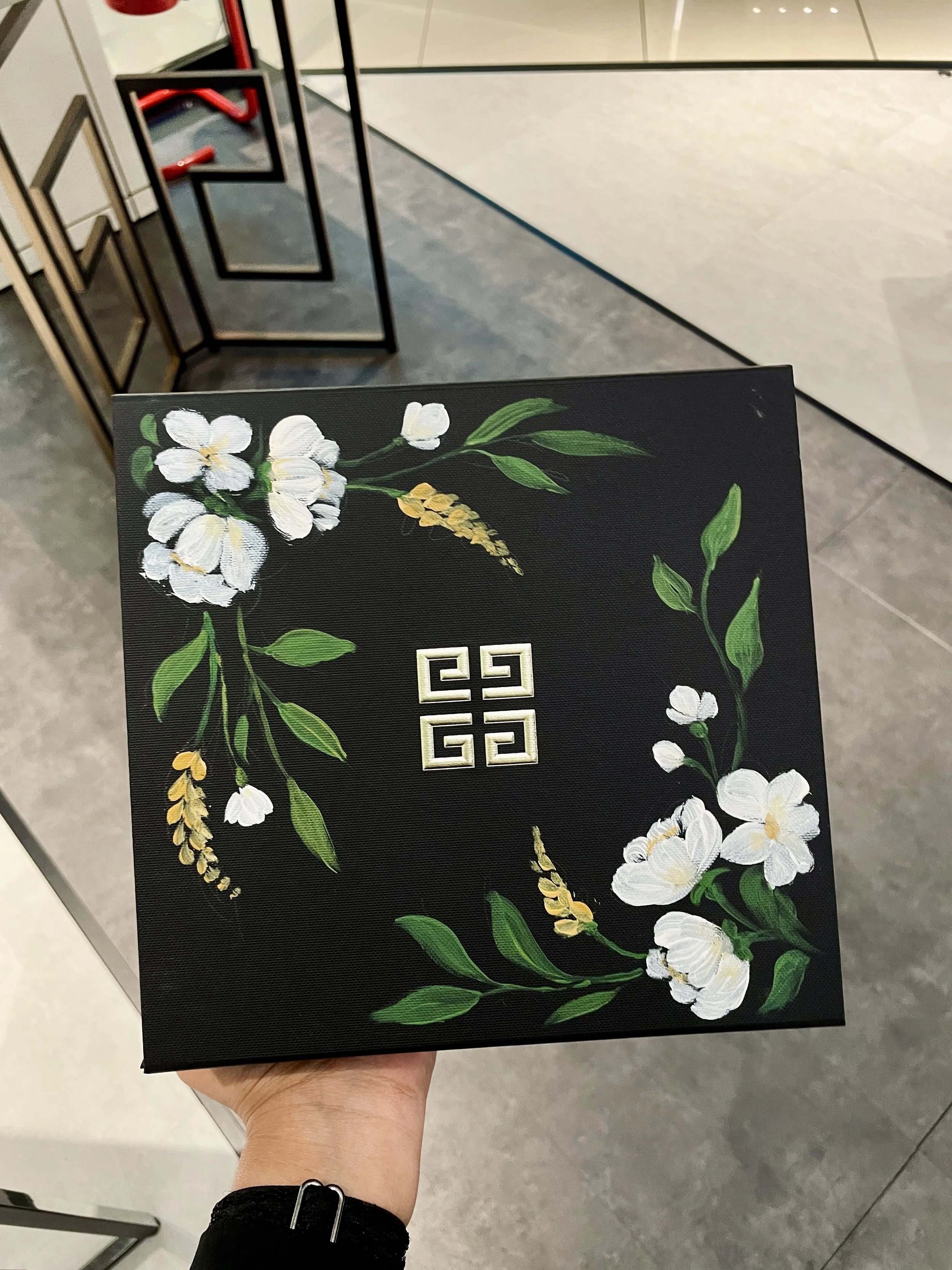 A person holding a black rectangular item with white flowers and green leaves illustrations, and a small gold-colored symbol in the center, in an indoor setting with tiled floors and a metal display stand in the background.
