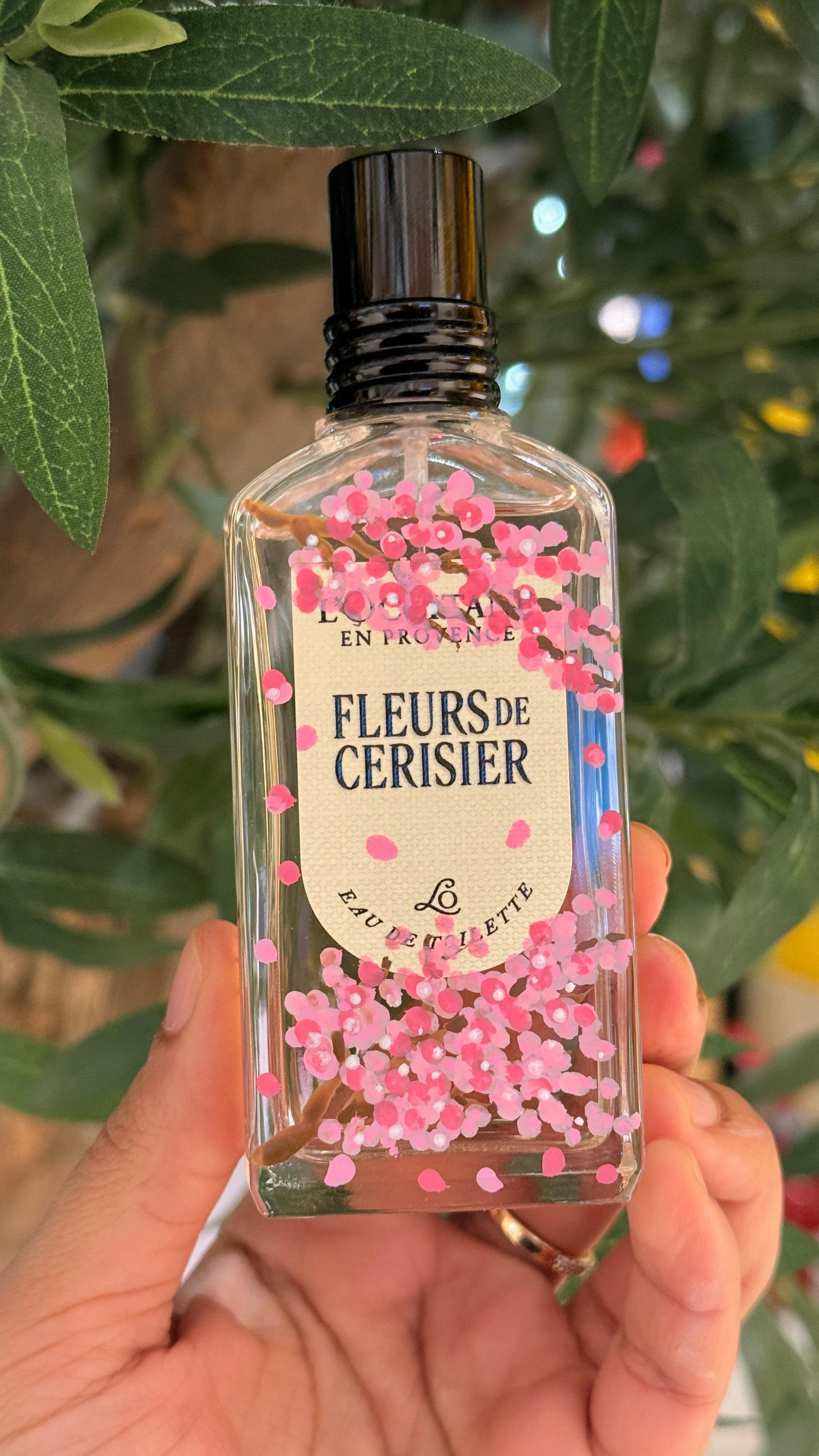 A hand holding a clear rectangular perfume bottle with pink cherry blossom petals inside. The label reads "Fleurs de Cerisier" and "Eau de Toilette". Green leaves are visible in the background.
