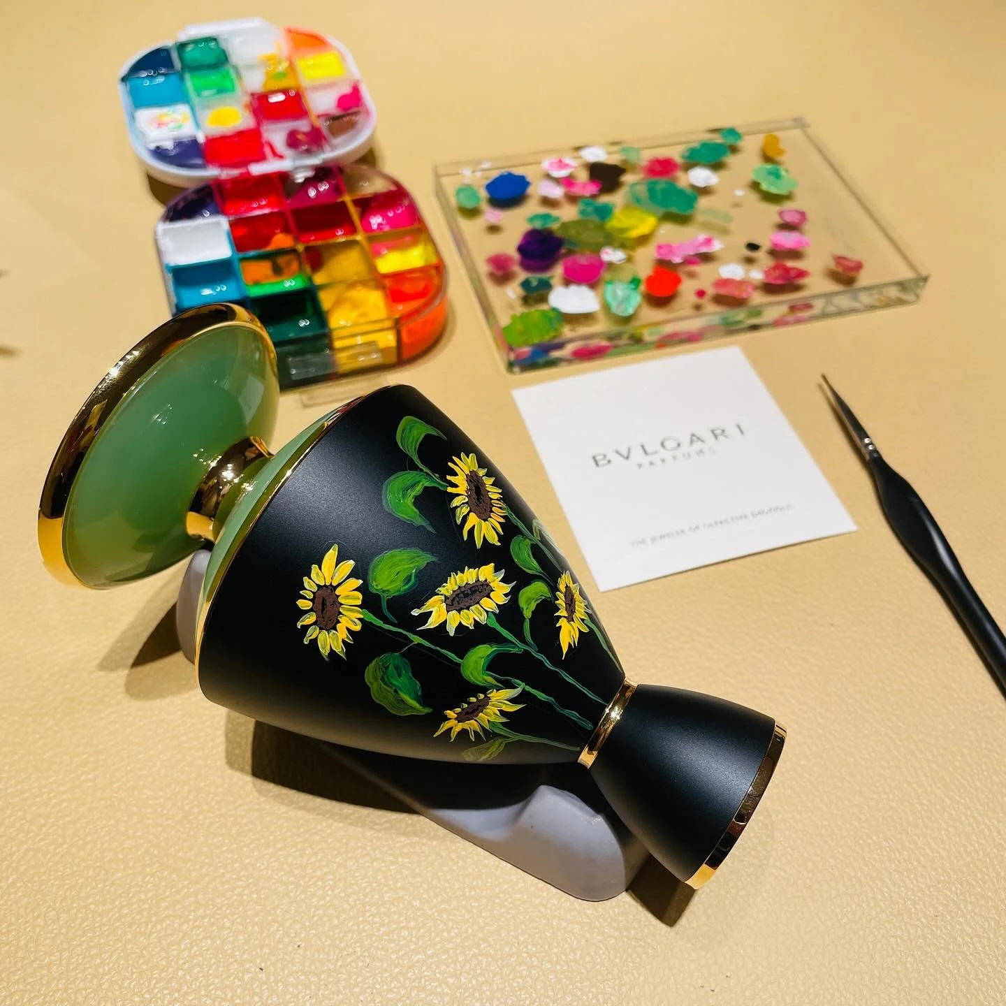 Decorative black perfume bottle with sunflower design, perfume set with colorful paint palette, and flower decorations on a beige surface.