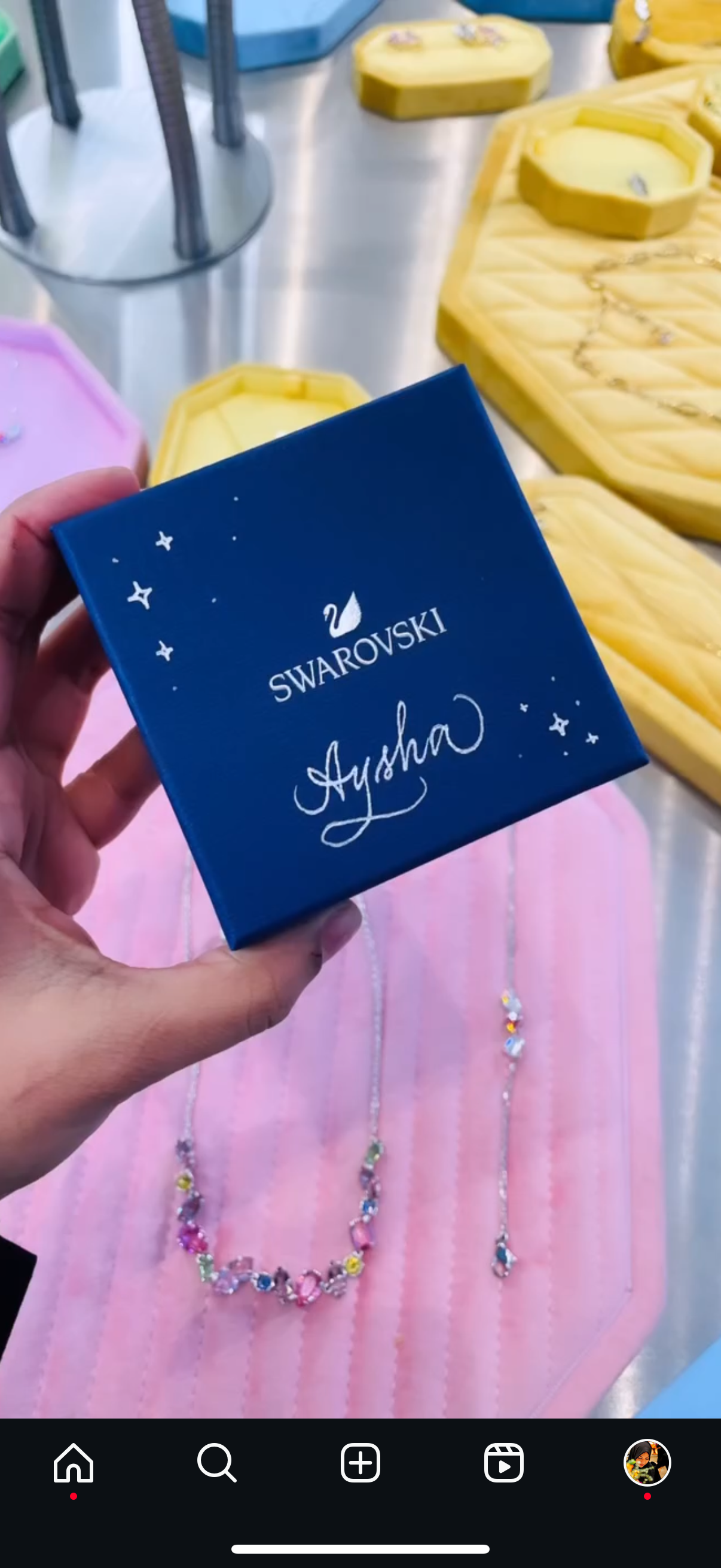 A hand holding a blue Swarovski box with the name Aysha on it, against a background with pink and blue objects, a necklace with colorful stones, and yellow soap bars.