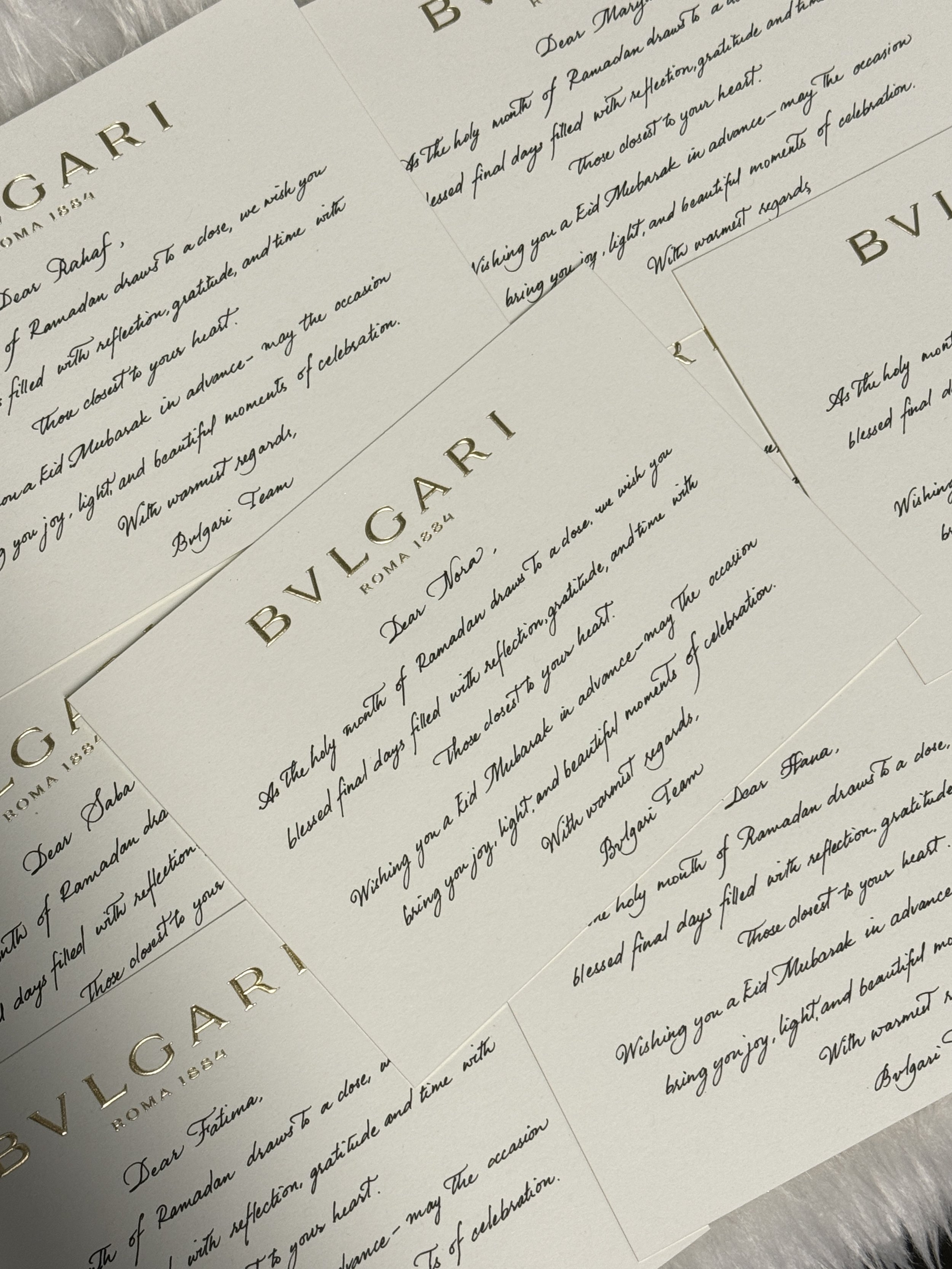 Close-up of several elegant letter-sized cards with the title 'BVLGARI' and handwritten-style text inside, partially overlapping each other, on a textured surface.