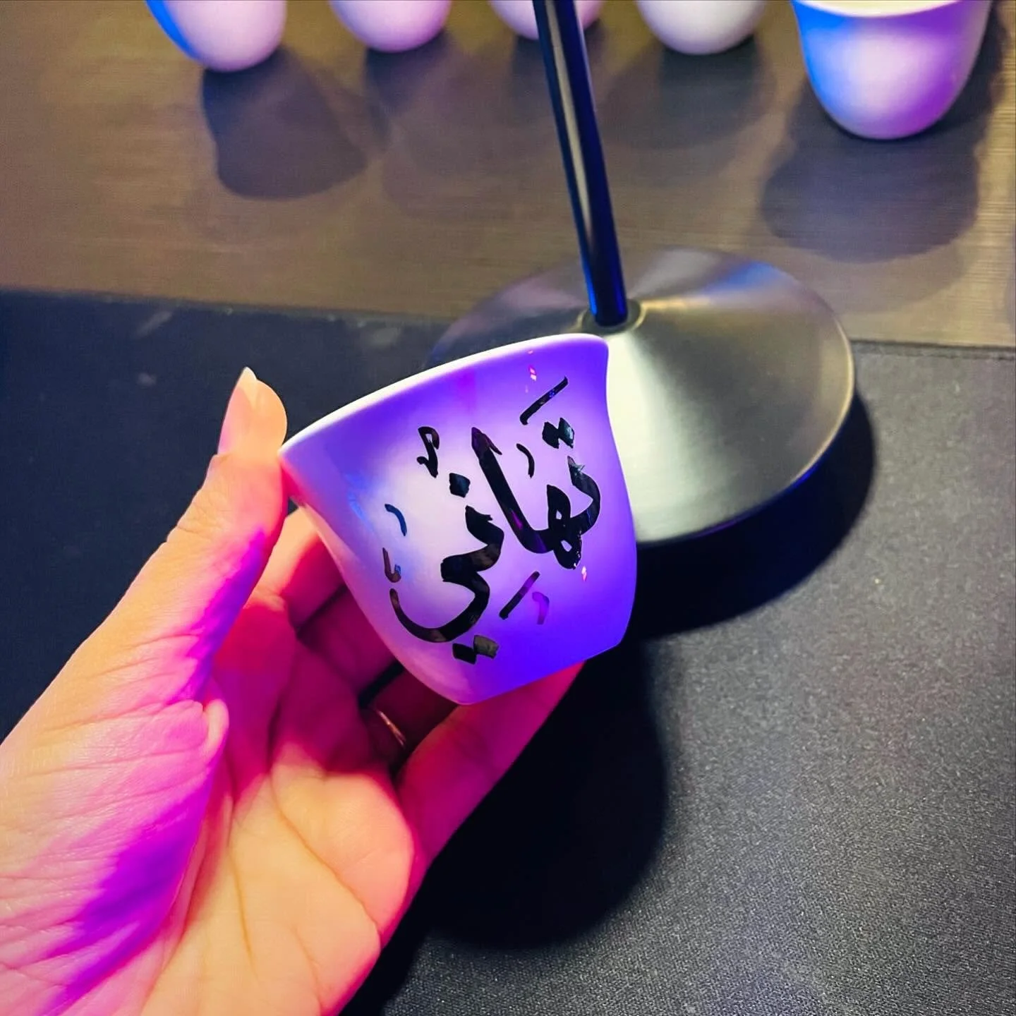 A person holding a white cup with black Arabic writing on it, illuminated by purple and pink lighting. The cup is tilted, and in the background, there are several metallic cups or bowls on a dark surface.
