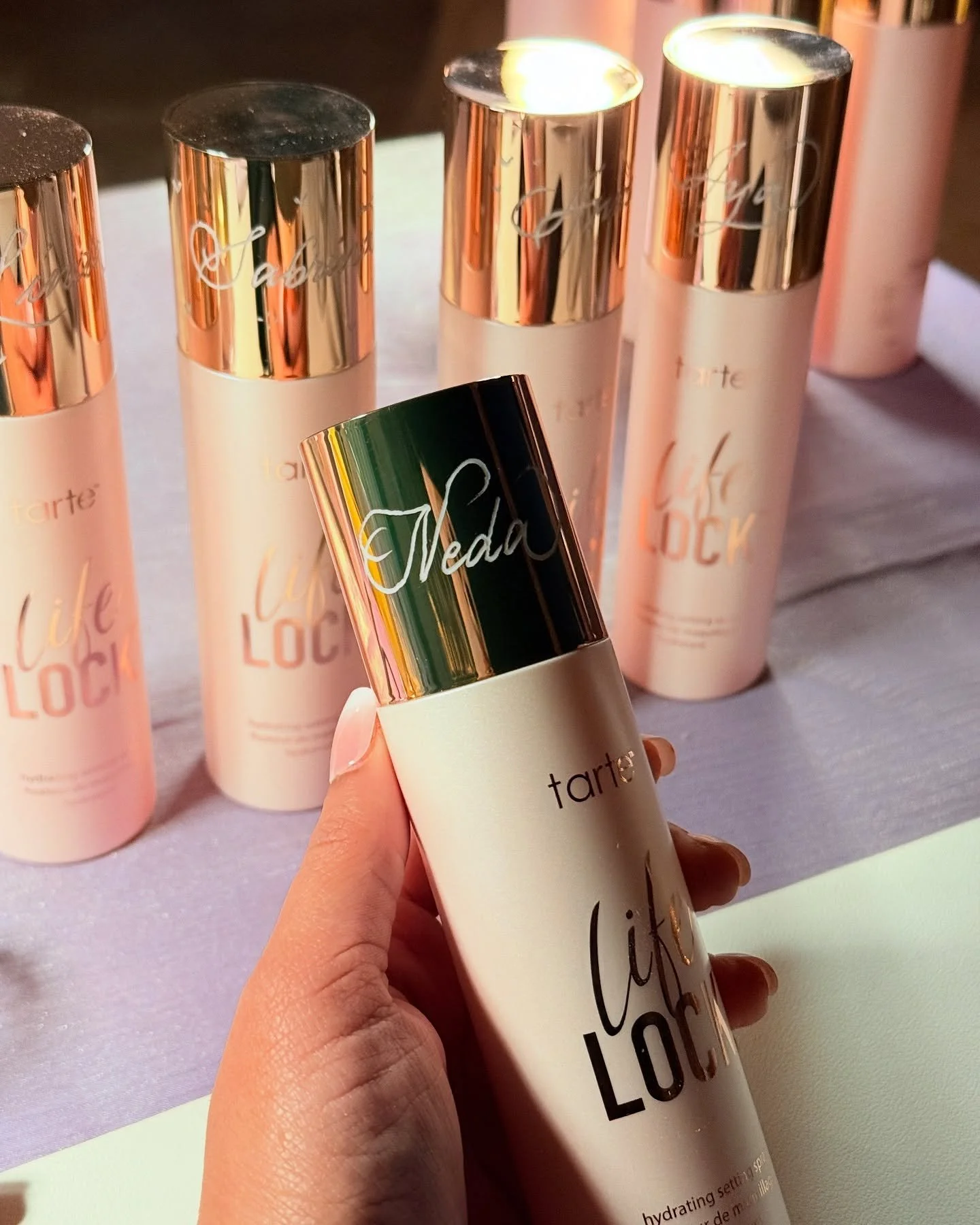 Close-up of a person holding a white bottle with a silver cap, labeled 'tarte' and 'Lick' in black, with several similar bottles in the background on a pink surface.