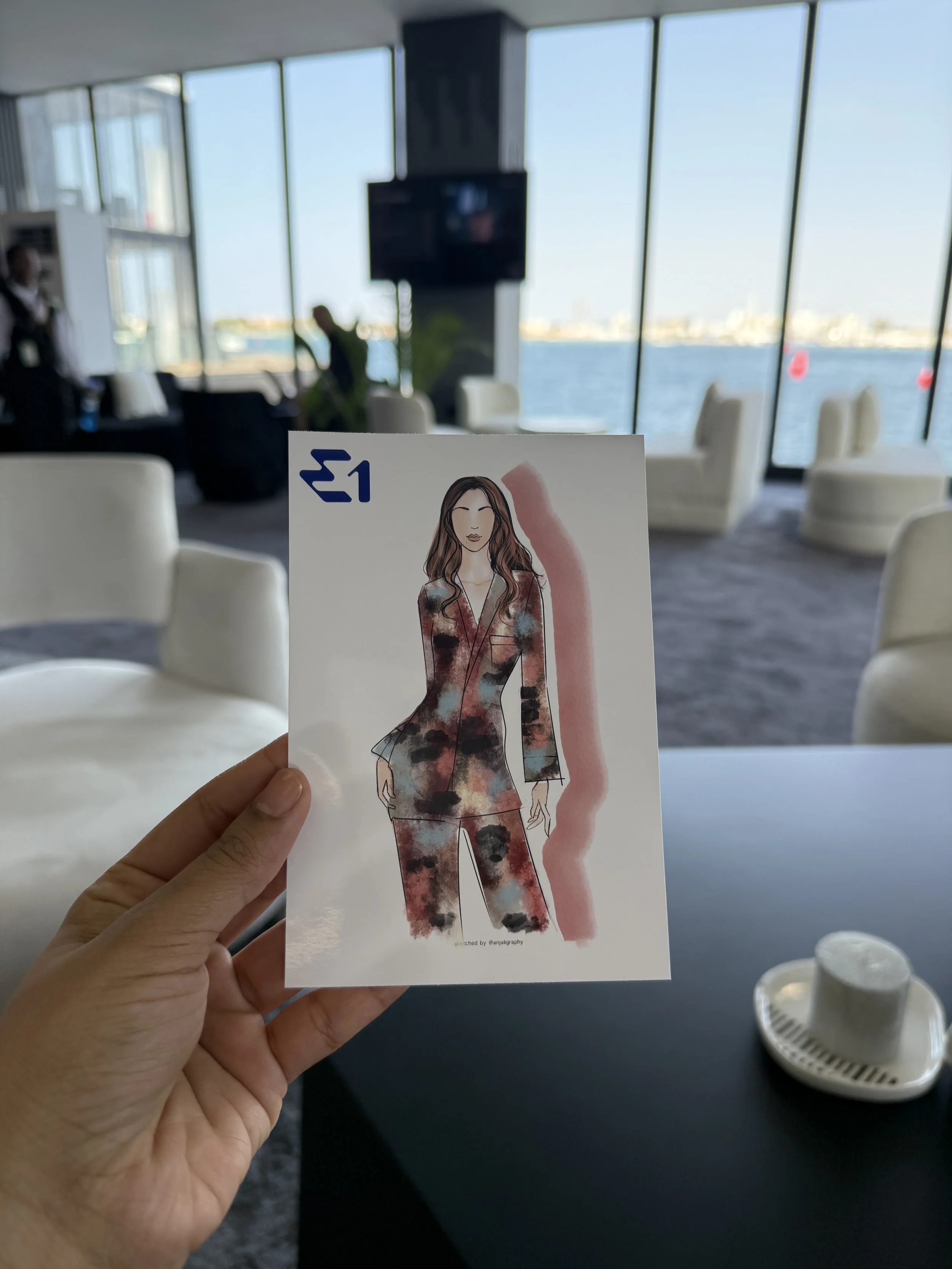 A hand holding a printed illustration of a woman with long wavy hair, wearing a floral jumpsuit, in front of a window overlooking water and cityscape on a sunny day in a lounge or waiting area.